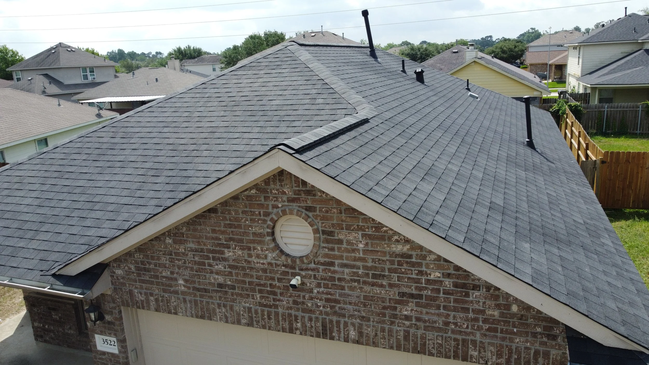 Aerial view of a house with a gray shingle roof, brick exterior, and white garage door in a neighborhood of similar houses with fenced yards and green lawns.