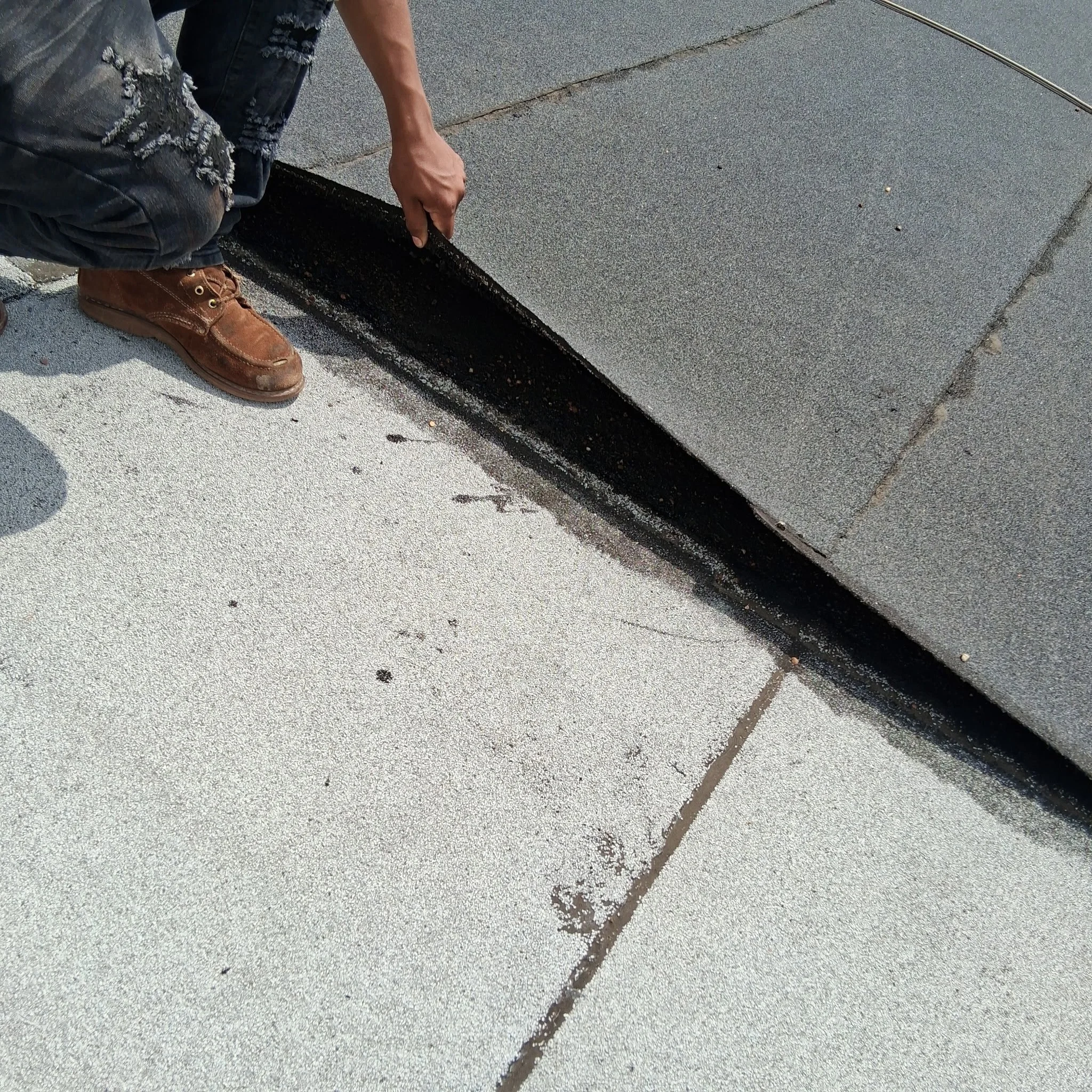 Person kneeling on concrete sidewalk, installing black waterproof membrane along the edge of a building's foundation with their right hand, wearing ripped jeans and brown work boots.