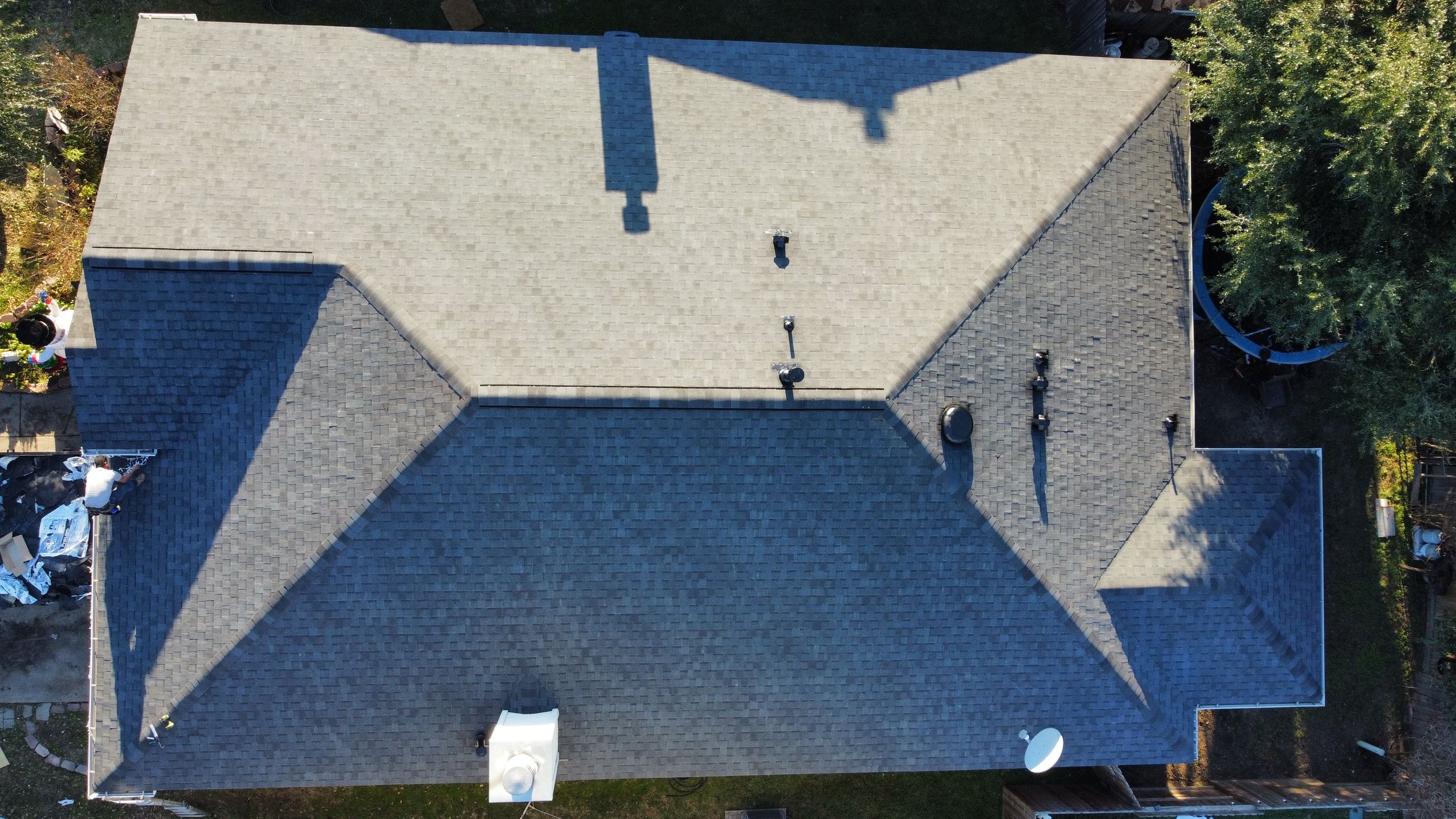 Aerial view of a house roof with multiple vents, chimneys, and HVAC units, surrounded by trees and a backyard.
