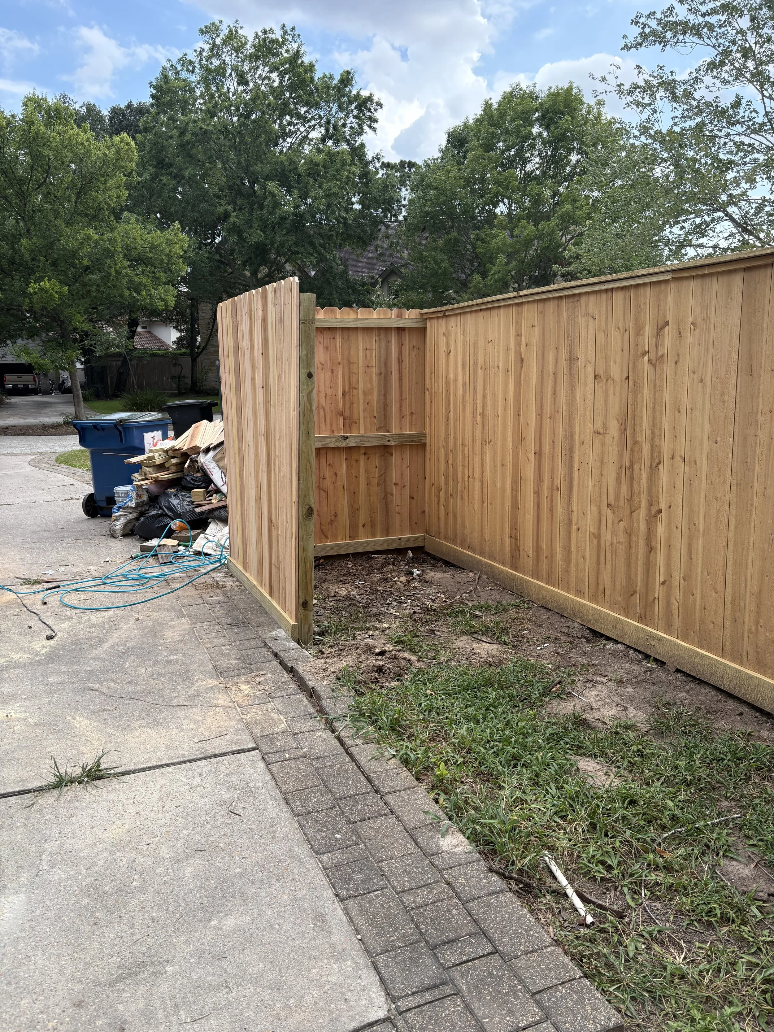 New wooden privacy fence being installed along a sidewalk. Construction tools, debris, and waste materials are visible on the ground.