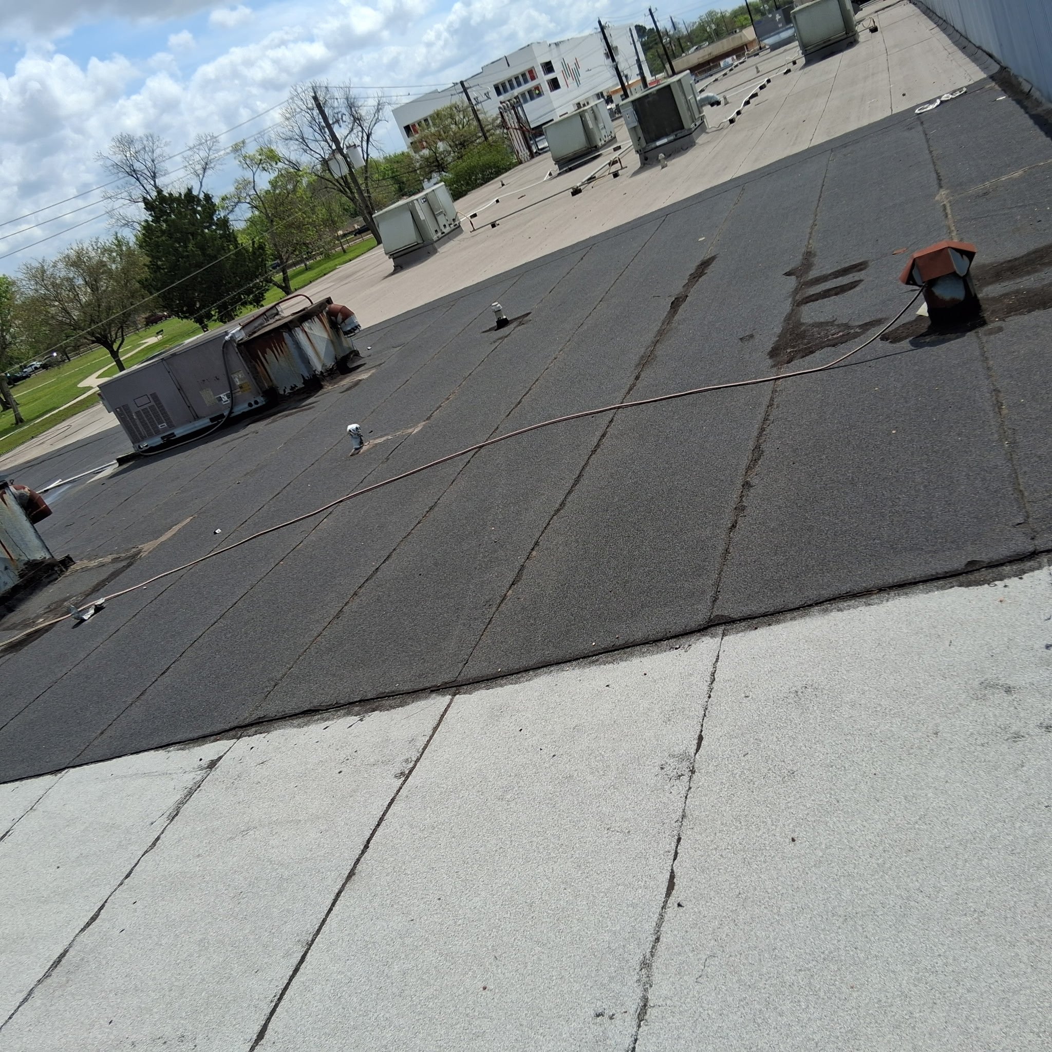 A flat rooftop with damaged black asphalt shingles, HVAC units, gutter, and vent pipes, with a park and blue sky with clouds in the background.