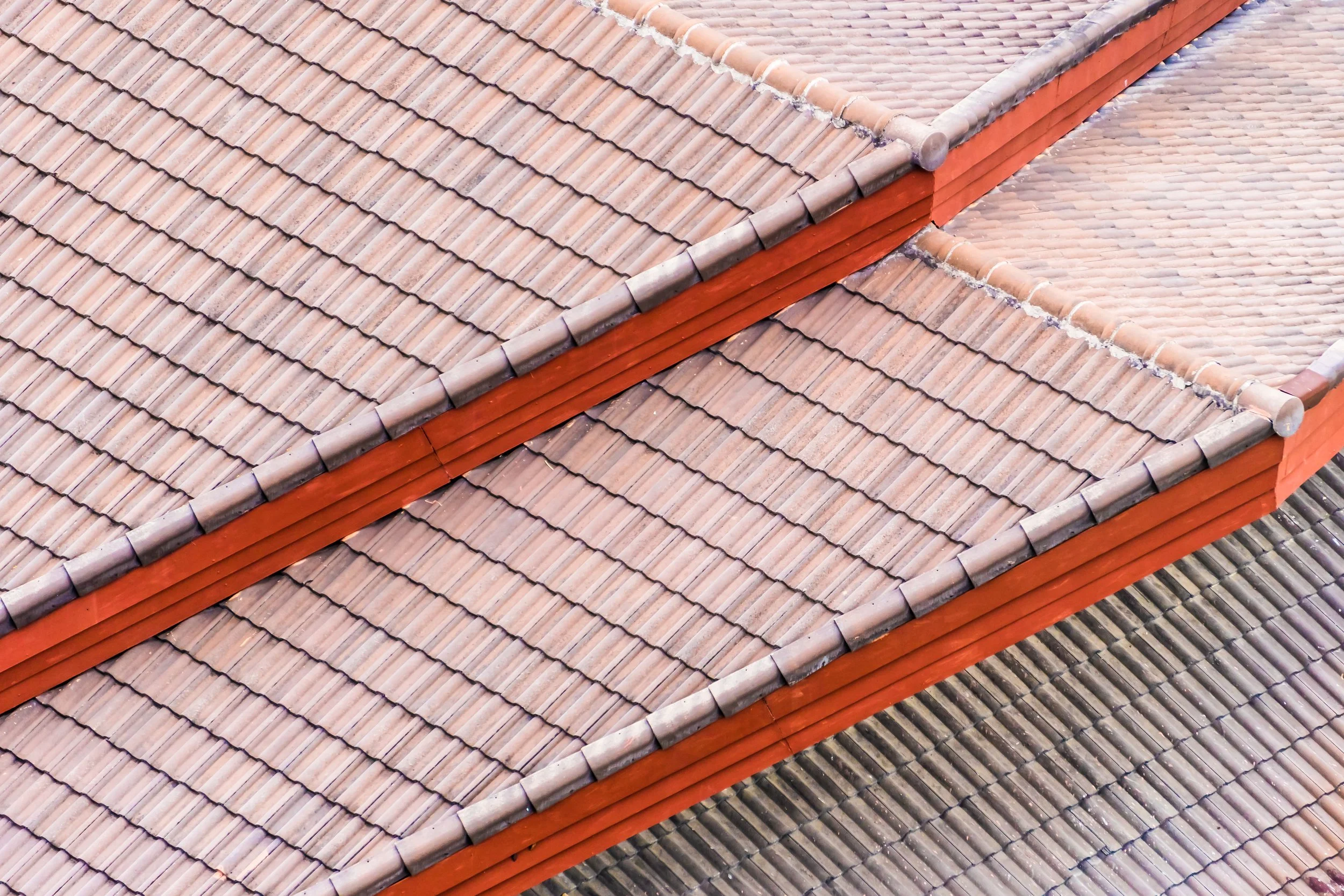 Close-up aerial view of tiled roof with reddish-brown ridge tiles and different shades of roof tiles in pink and gray.