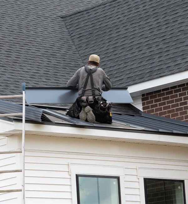 A construction worker installing or repairing metal roofing on a residential house. The worker is kneeling on the roof and wearing a safety belt and helmet.