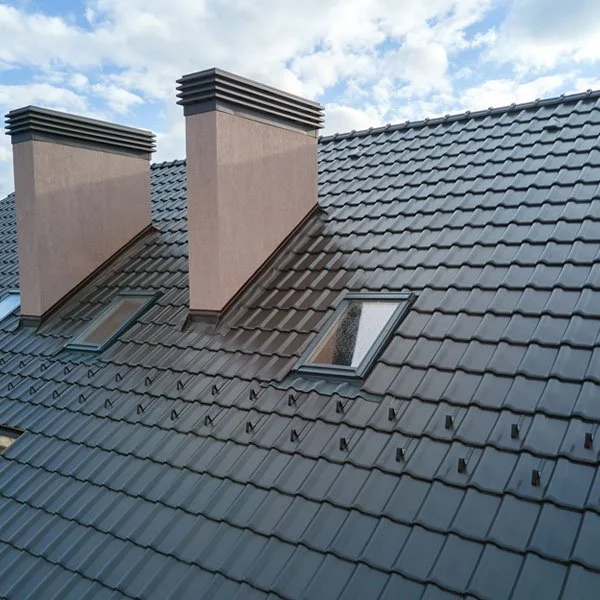 Close-up of a rooftop with dark gray ceramic tiles, two pink chimney stacks, and two skylight windows, under a partly cloudy sky.