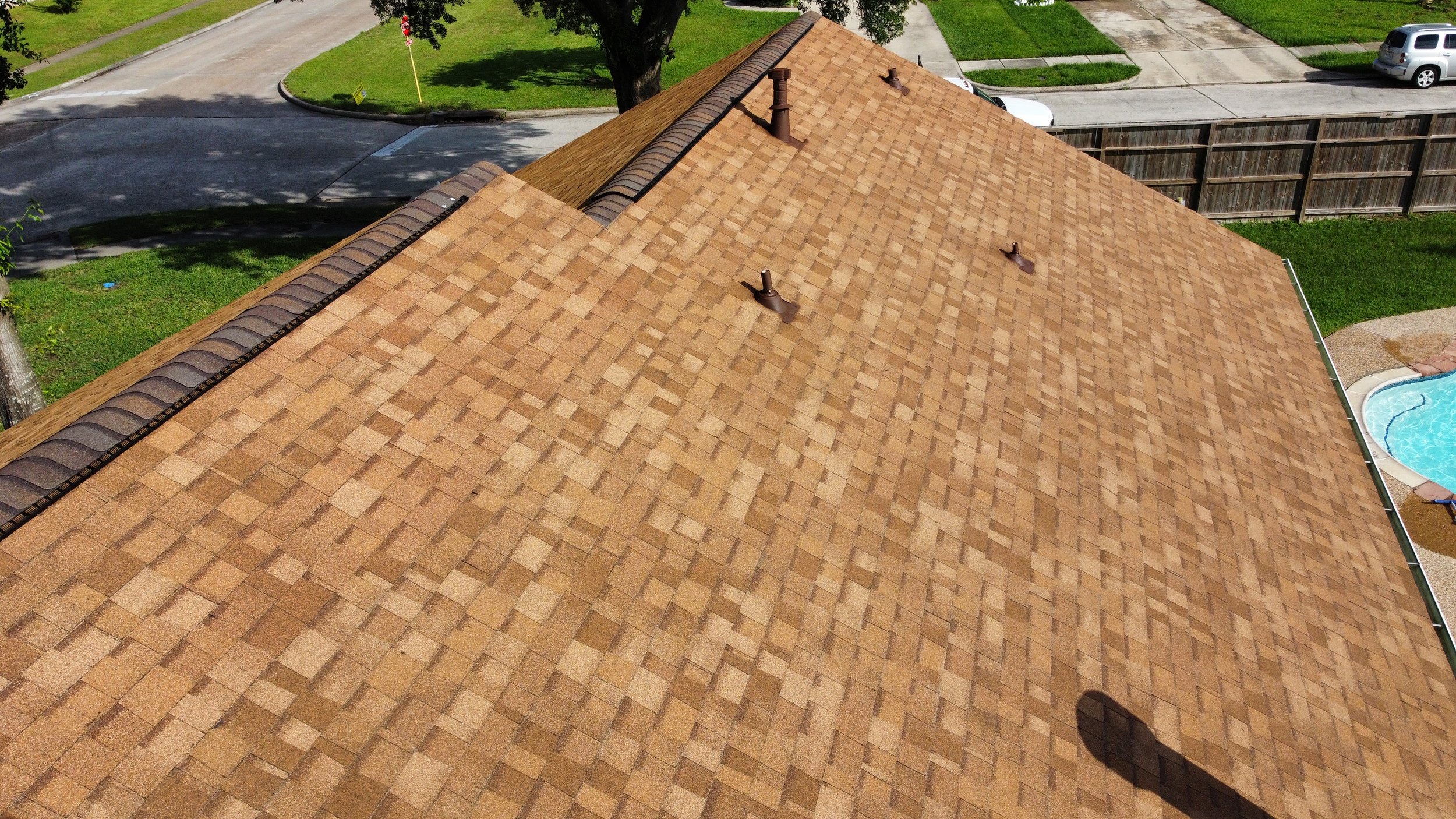 Aerial view of a house's brown shingle roof with small vents, surrounded by a green lawn, a wooden fence, a swimming pool, and nearby roads with parked cars.