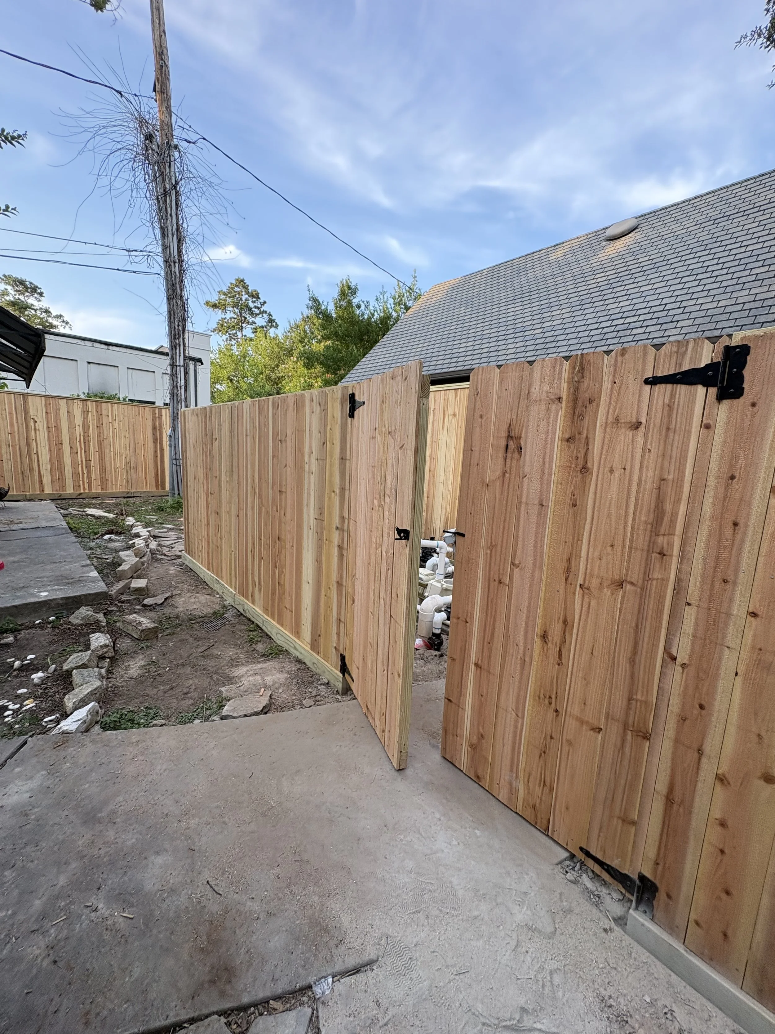 A wooden fence with black hinges partially open, revealing pipes underneath. The ground is dirt with some rocks and debris, and in the background, there is a building with a gray shingled roof and trees behind it. A power line pole is visible in the 