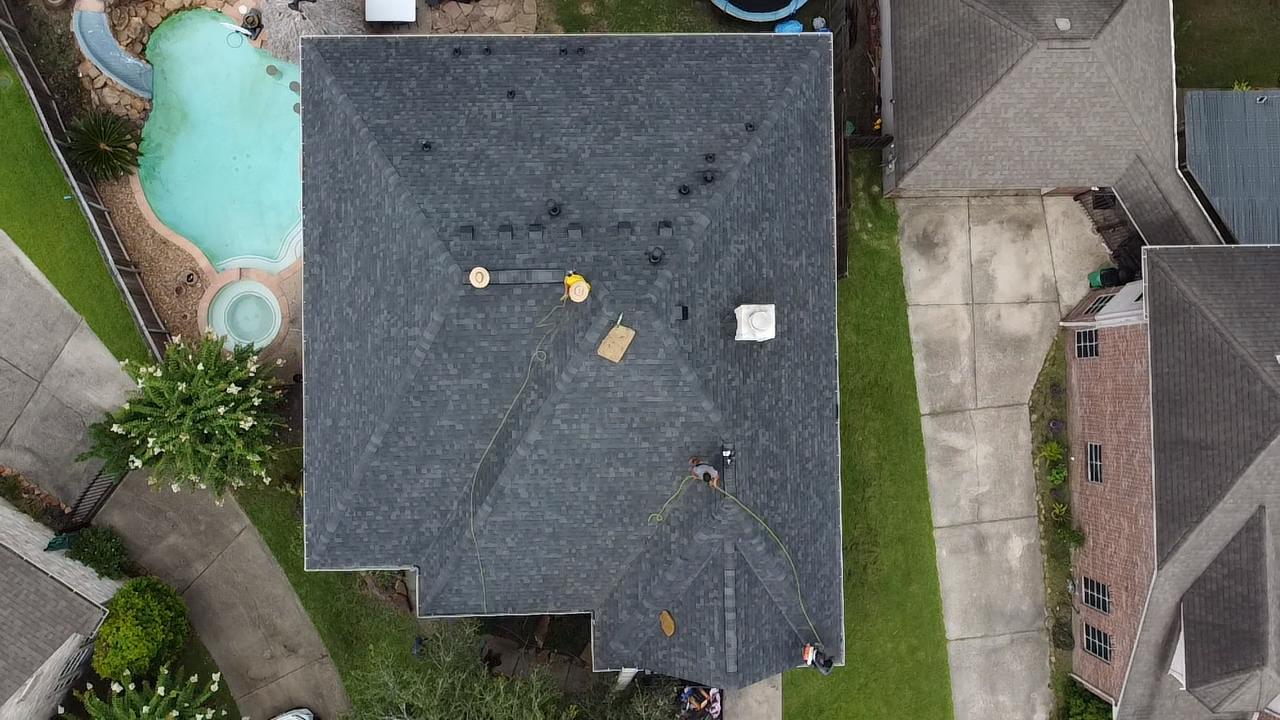 An aerial view of a house roof with three workers installing or repairing roof shingles. Surrounding the house are neighboring houses, a backyard with a pool, and a backyard with a green lawn.