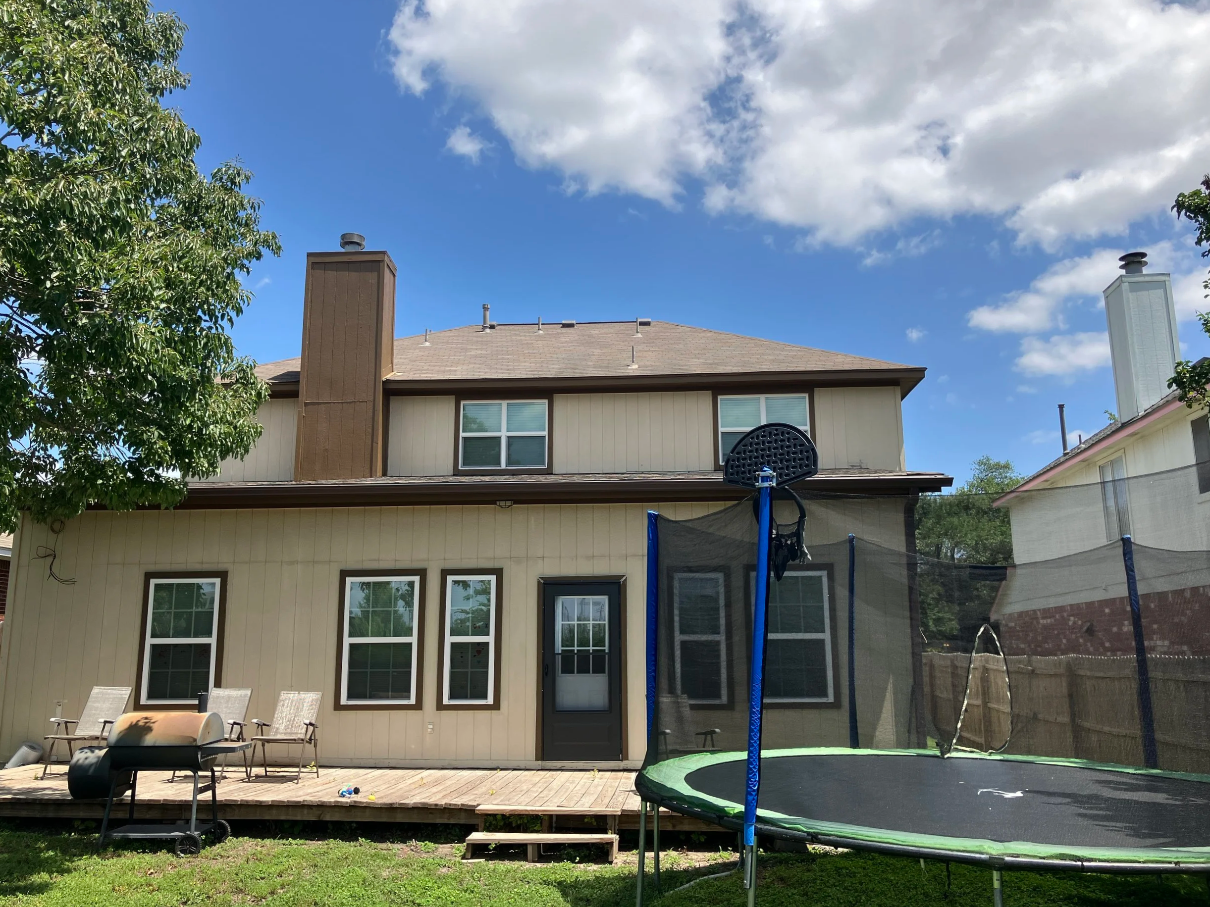 Backyard patio with outdoor chairs, a trampoline, a chair, and a house with windows and a sliding door under a mostly cloudy sky.