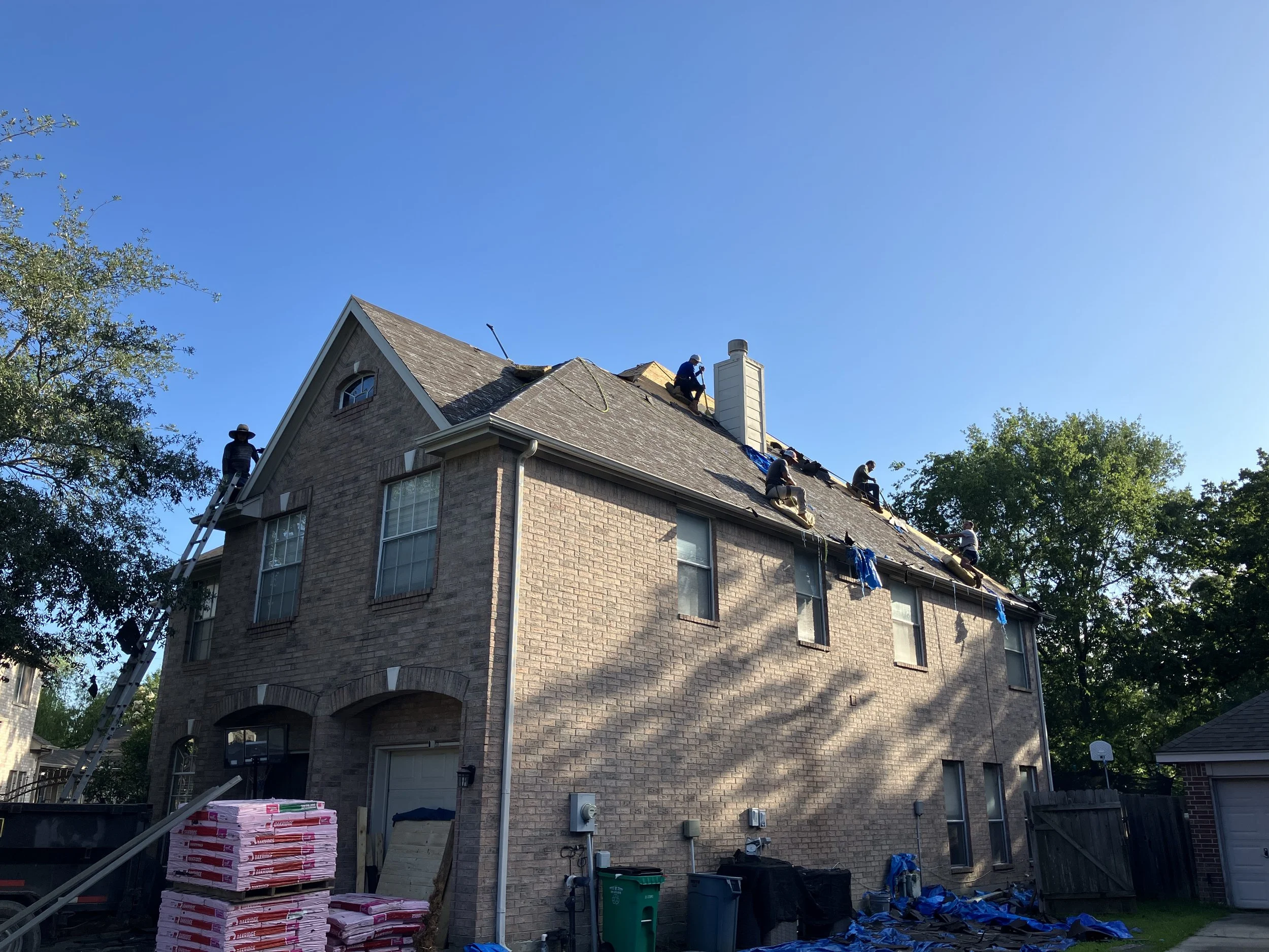 Workers repairing the roof of a multi-story brick house under a clear blue sky, with construction materials and tools around the yard.