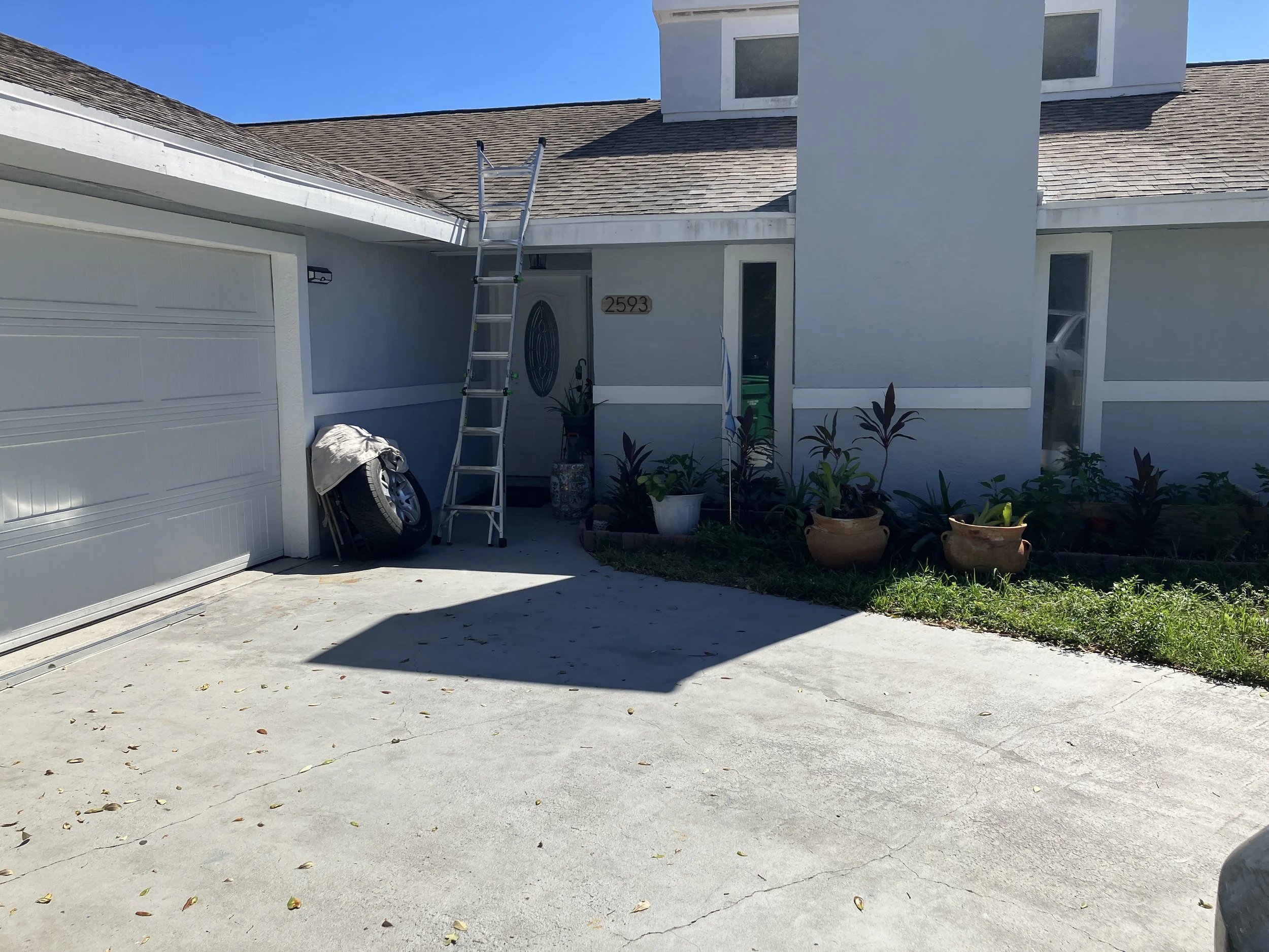 Front of a residential house with a driveway and garden. A silver ladder is leaning against the house under the roof overhang. There are labeled house numbers 2593 near the front door. Several potted plants and a tire are near the entrance, with a ga
