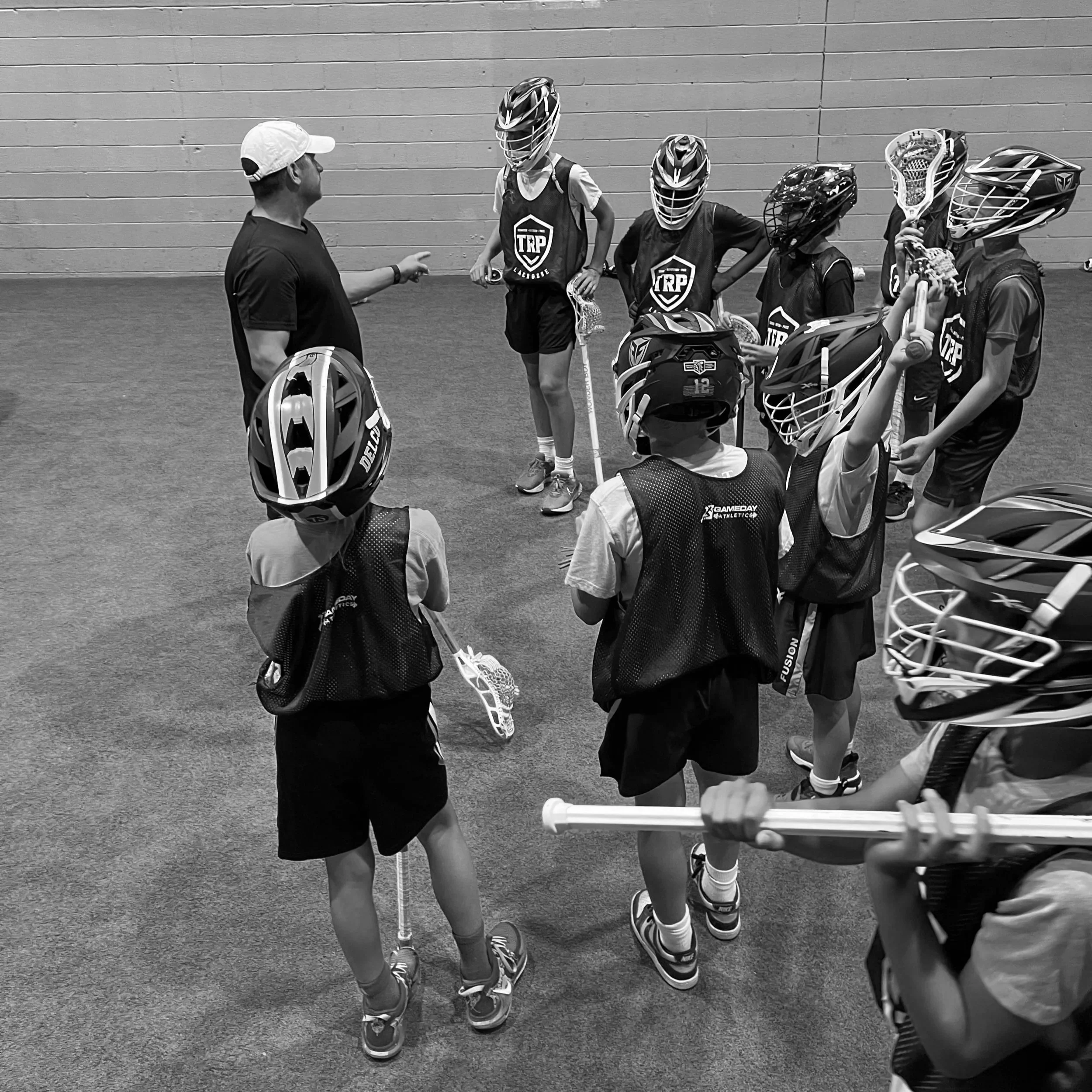 A coach or instructor talking to a group of young lacrosse players in an indoor sports facility. The players are wearing helmets and athletic gear, and are holding lacrosse sticks.