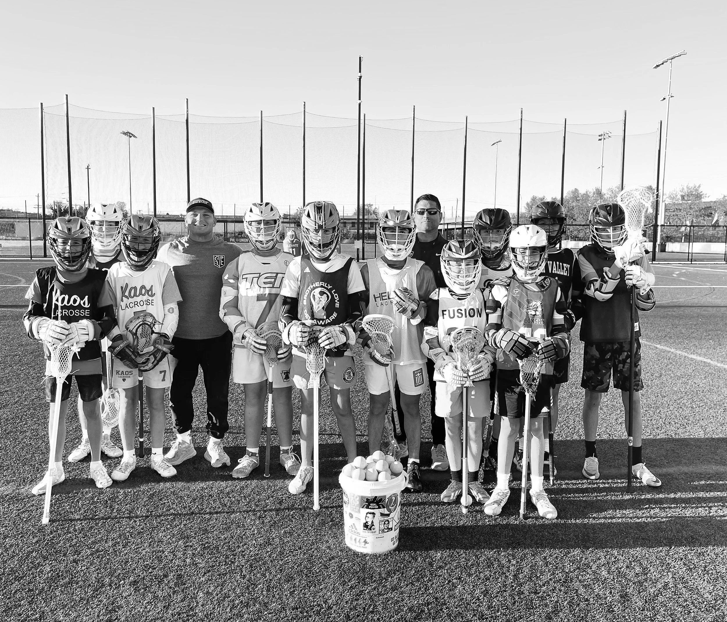 A group of young lacrosse players and their coaches posing on a lacrosse field with a bucket of lacrosse balls. The players are dressed in team jerseys and gear, standing in front of a tall fence and lighting poles. The image is in black and white.