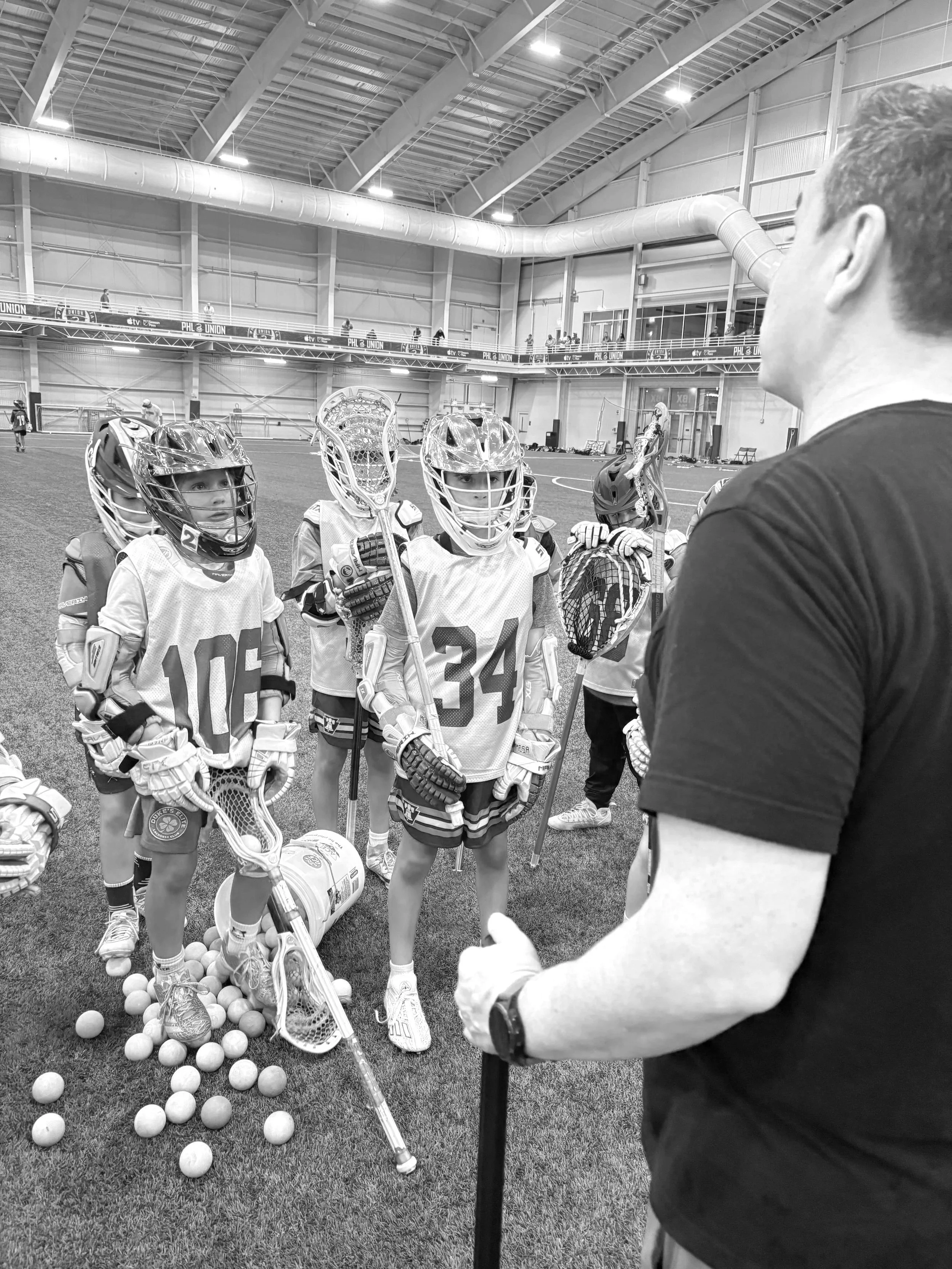 Young boys wearing lacrosse helmets and gear listening to coach inside an indoor lacrosse facility, with lacrosse balls on the ground.