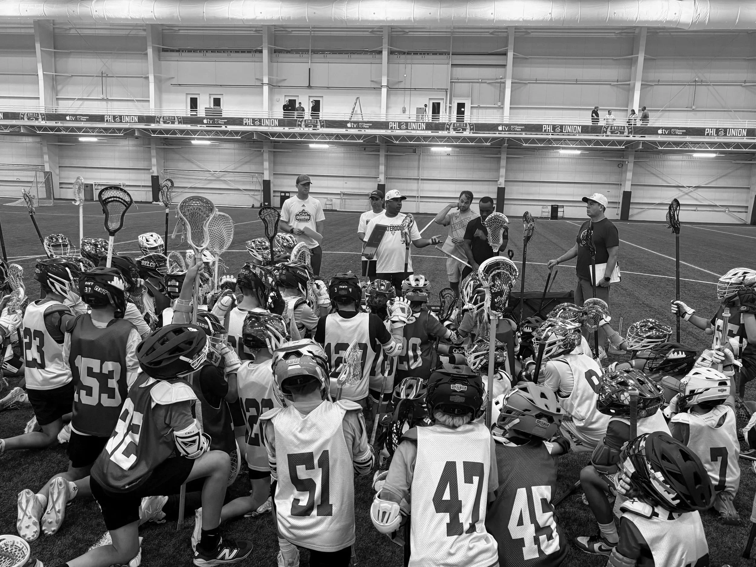 Indoor lacrosse practice with players and helmets, some holding sticks, in a large field with a high ceiling.