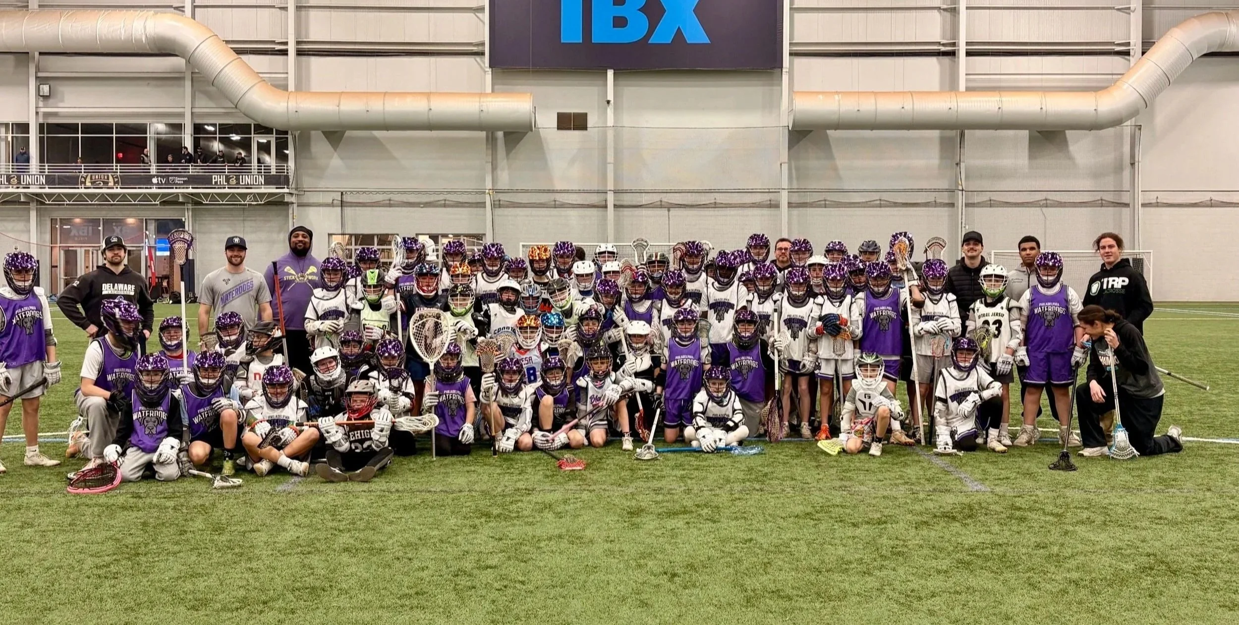 Lacrosse team with players and coaches posing on an indoor field, wearing uniforms and helmets, with a large IBX sign on the wall behind them.