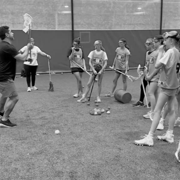 Girls playing indoor lacrosse practice with coach demonstrating technique, cones, and lacrosse balls on turf field.