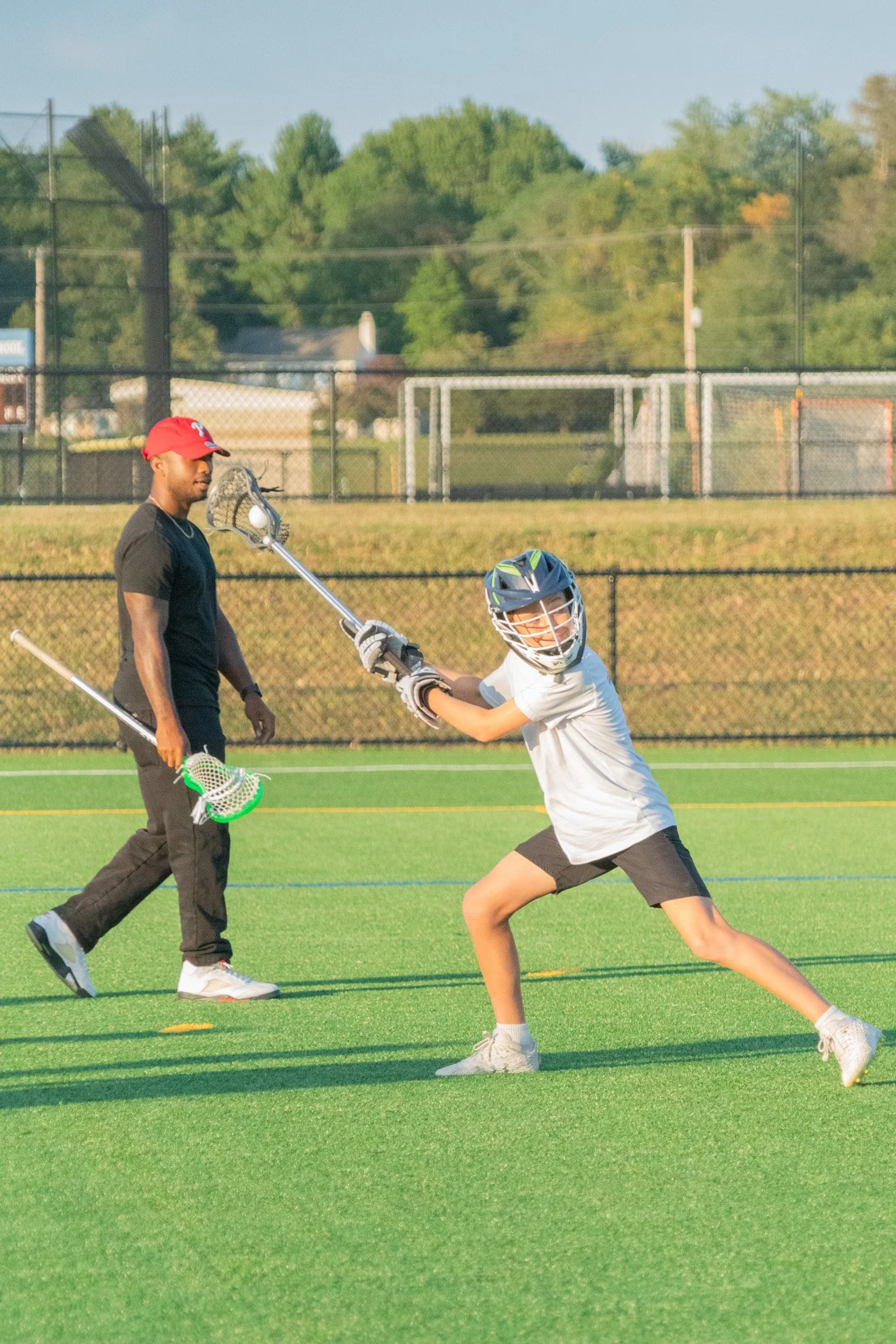 A boy wearing a helmet and white shirt playing lacrosse on a field, with an adult man standing nearby holding a lacrosse stick.
