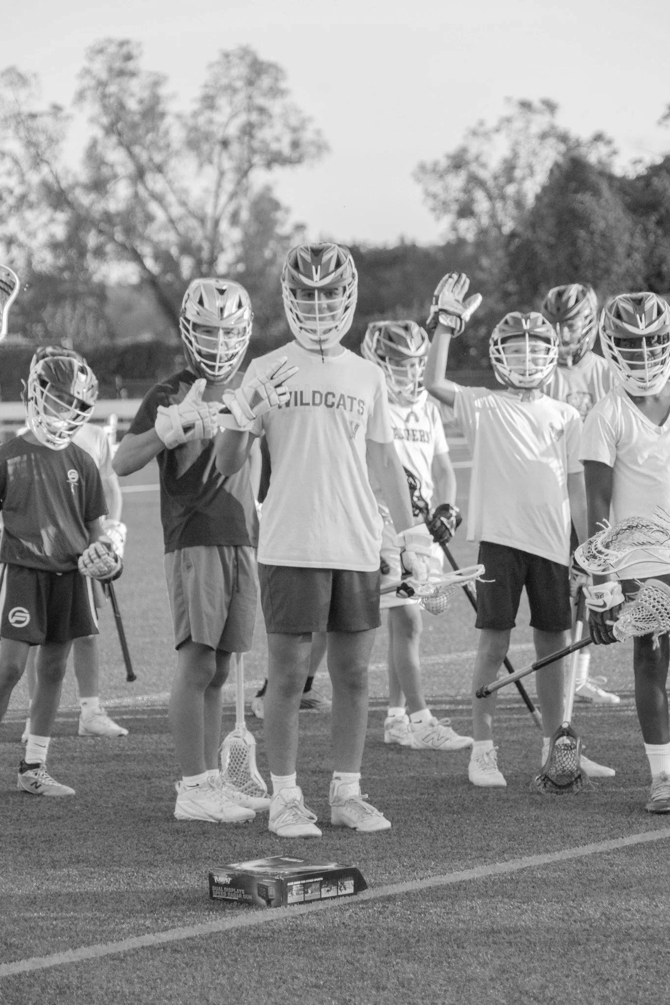 A group of young lacrosse players wearing helmets, gloves, and sports gear, standing on a field and waving at the camera.
