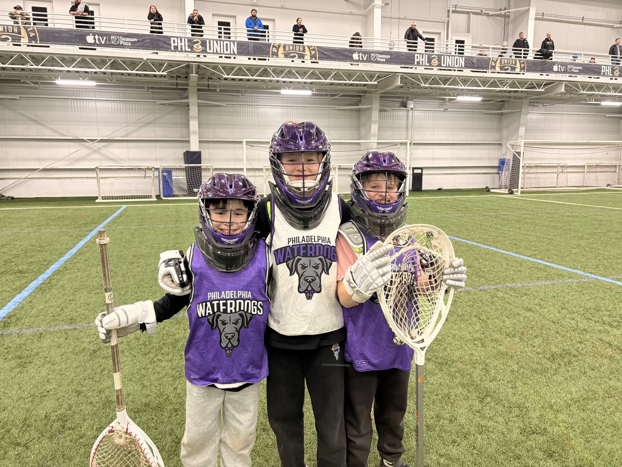 Three children and one adult on an indoor lacrosse field dressed in purple and black lacrosse gear, holding lacrosse sticks, smiling, with the children wearing jerseys that say 'Philadelphia Waterdogs' and a dog logo.