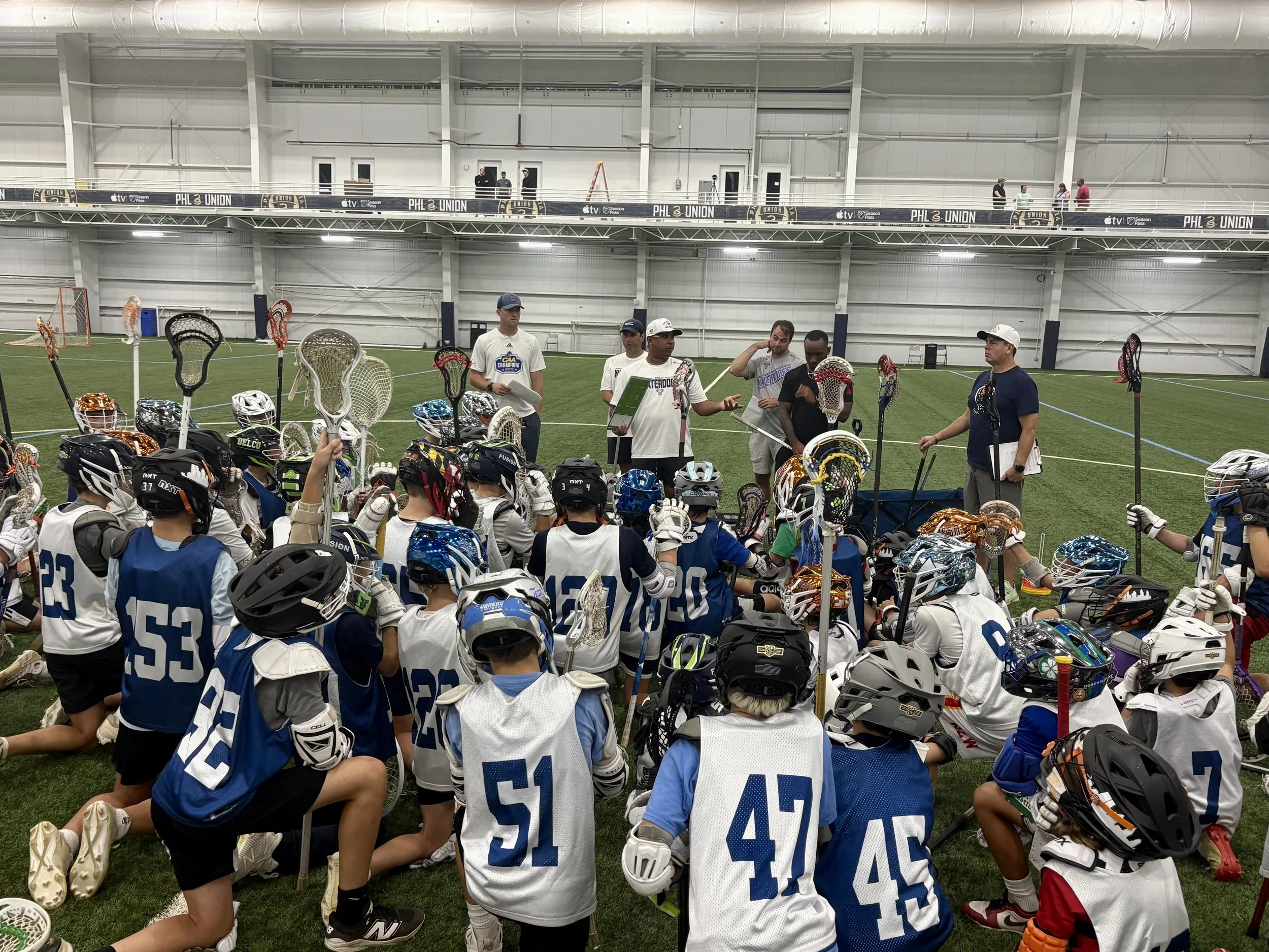 A large group of young lacrosse players wearing helmets and jerseys gathered on an indoor field listening to coaches during a team meeting or practice.