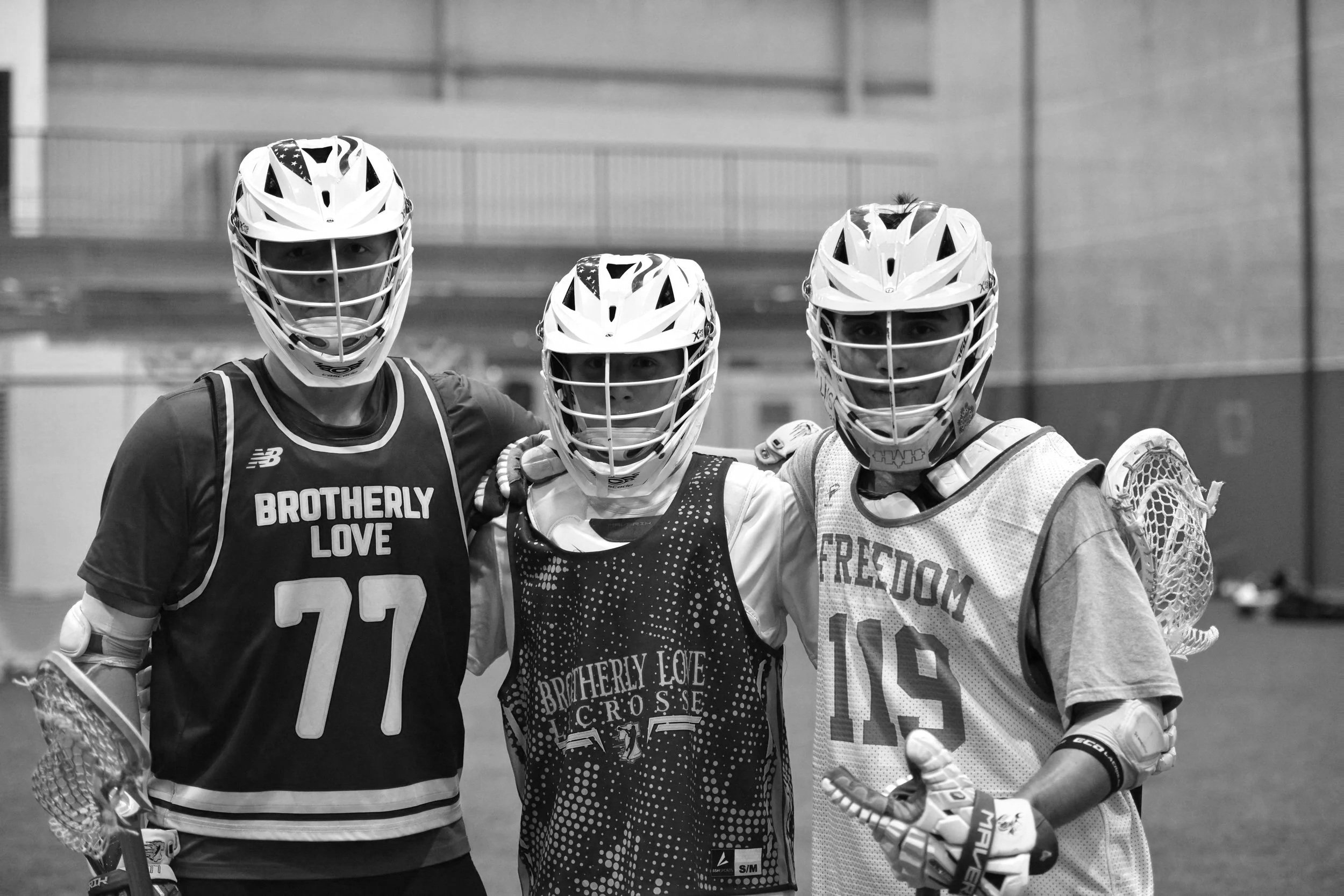 Three lacrosse players in helmets and protective gear standing together on an indoor field.