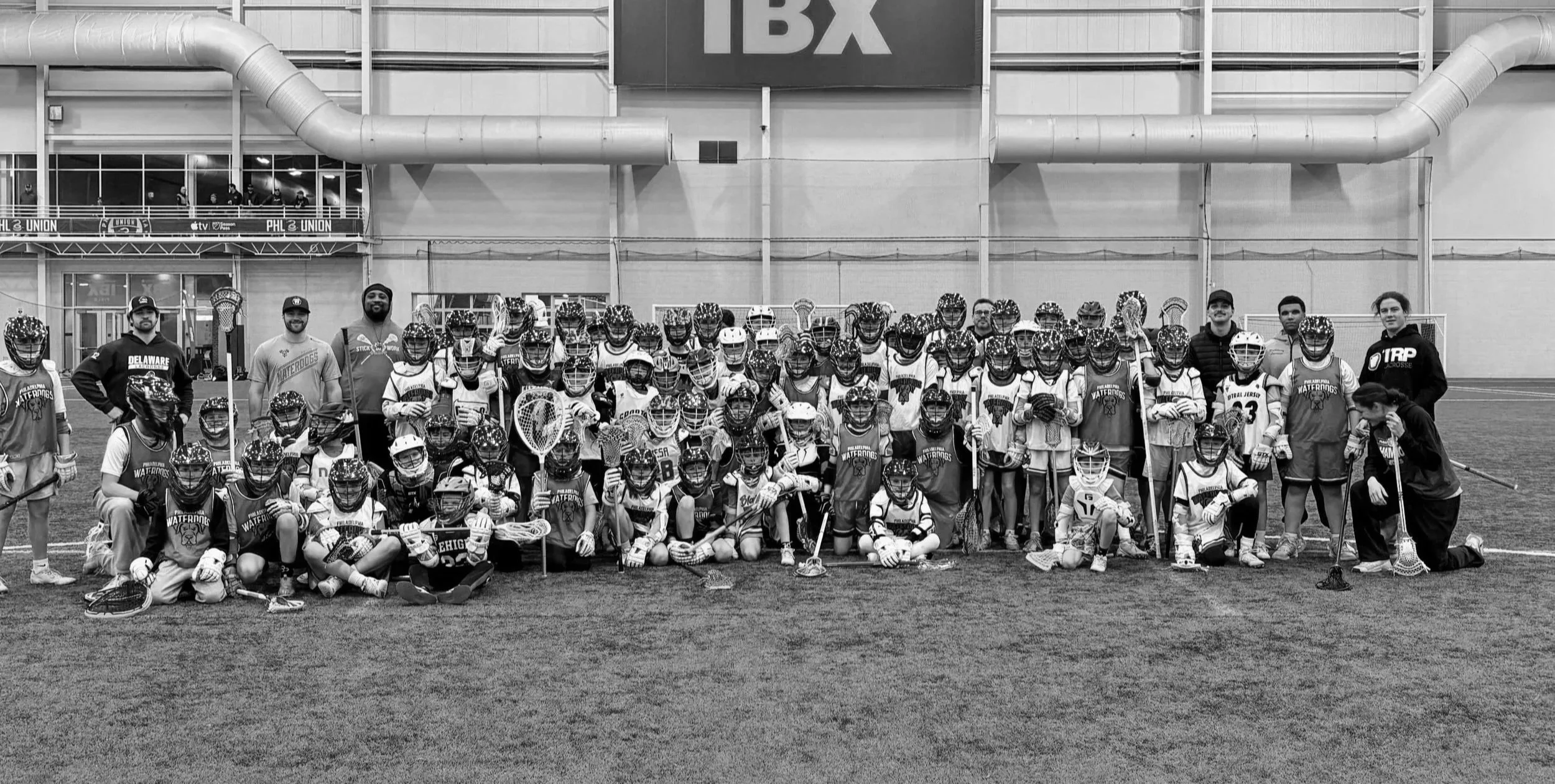 Black and white photo of a large group of young lacrosse players, coaches, and staff posing for a team photo on an indoor sports field, with some players kneeling and others standing, wearing uniforms and holding lacrosse sticks.