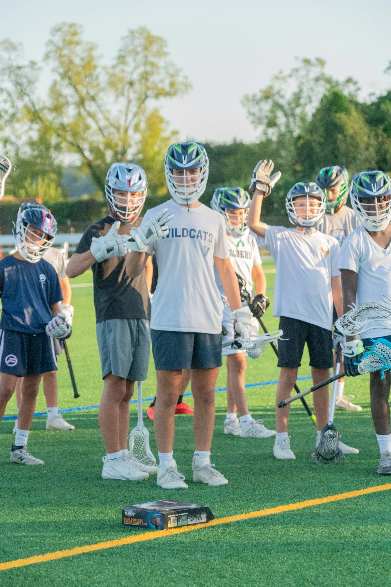 Group of young lacrosse players on a field, wearing helmets and sports gear, some holding lacrosse sticks, with trees in the background.