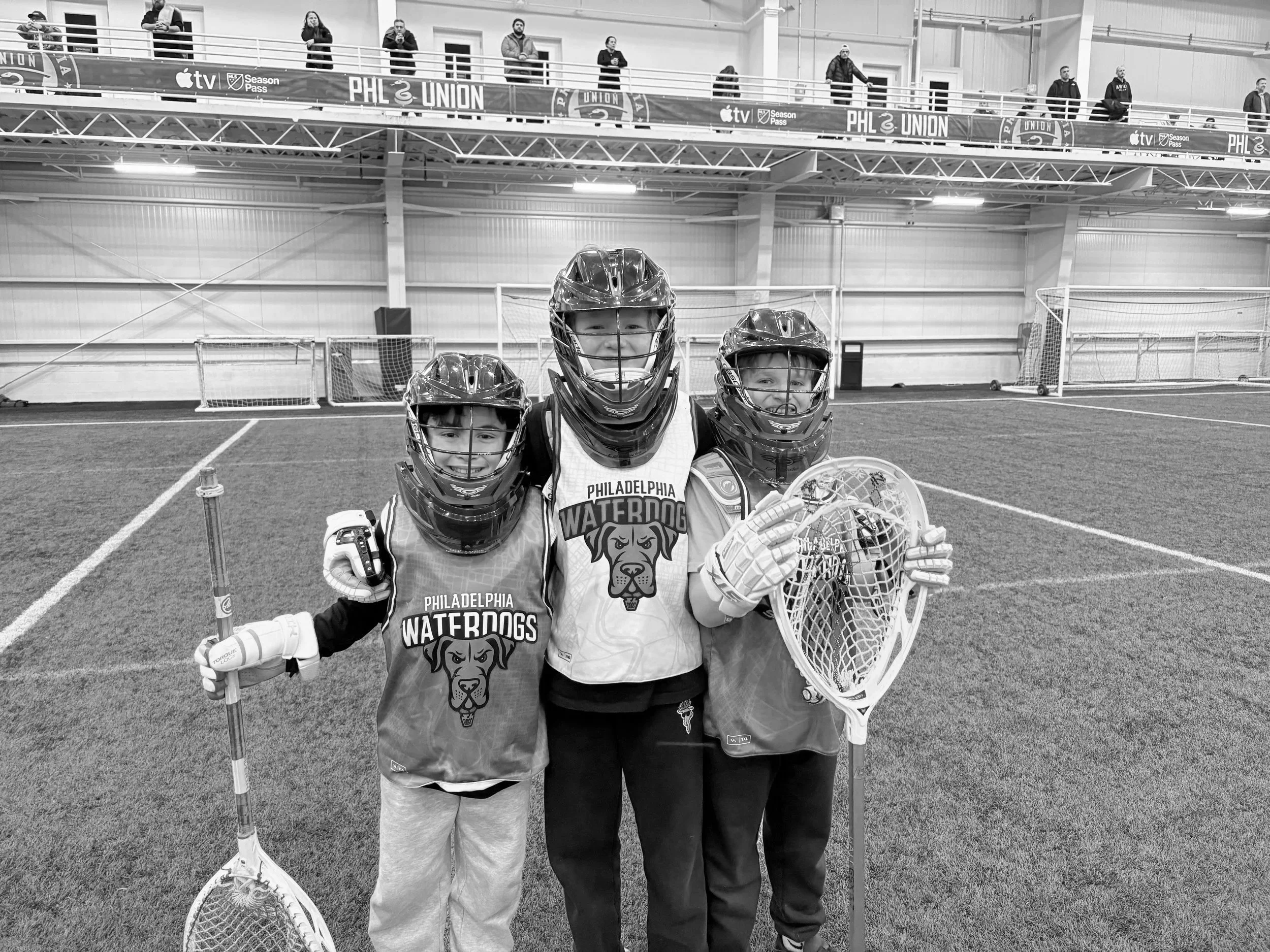 Three young children in hockey gear, smiling and posing together on an indoor sports field. Two are holding hockey sticks, and one is holding a goalie helmet, wearing matching jerseys with the words "Philadelphia Waterdogs" and a dog logo. They are standing in front of small hockey nets, with spectators watching from a balcony above.
