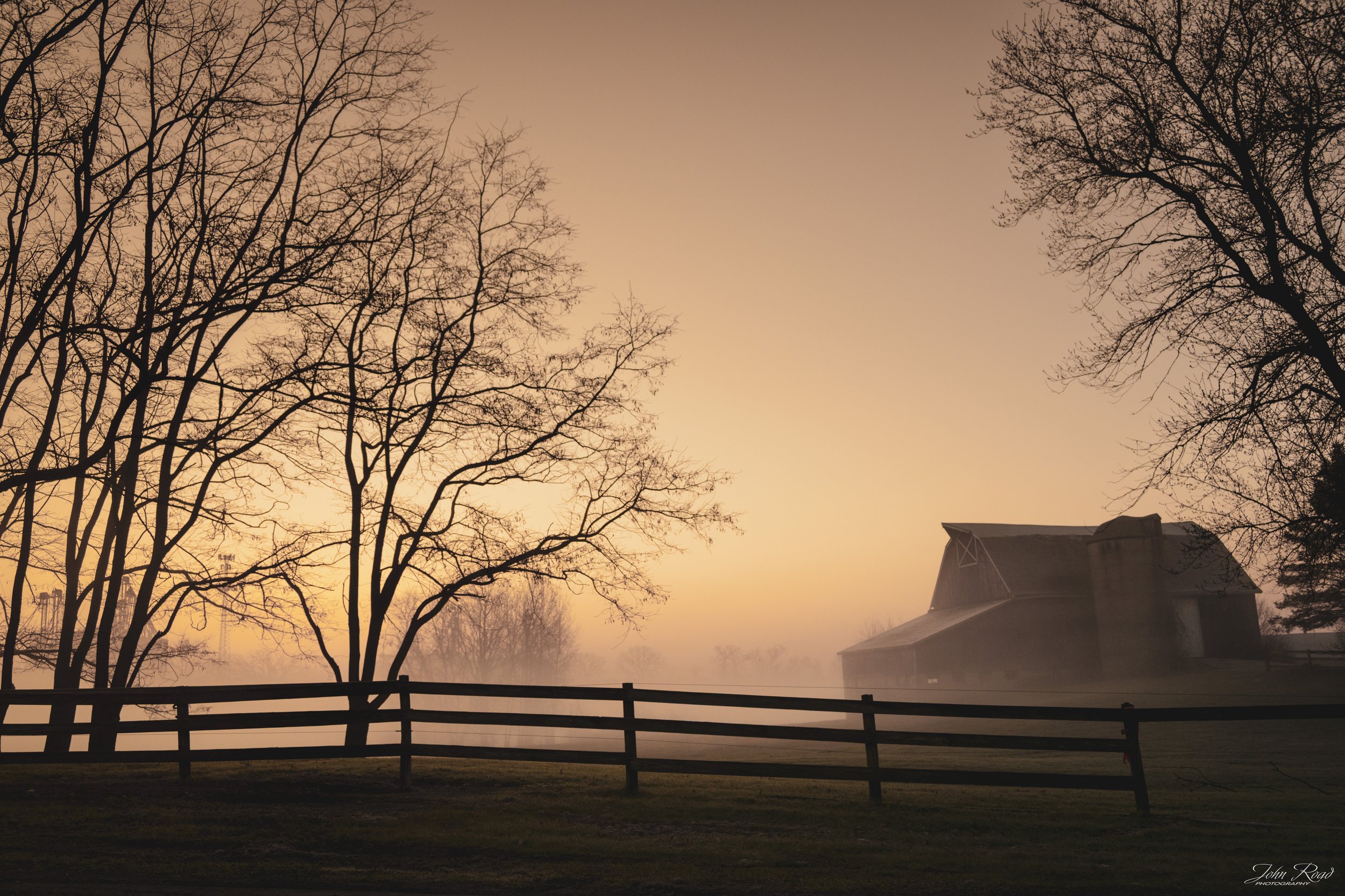 Foggy sunrise over a rural landscape with a barn, wooden fence, and bare trees in warm morning light