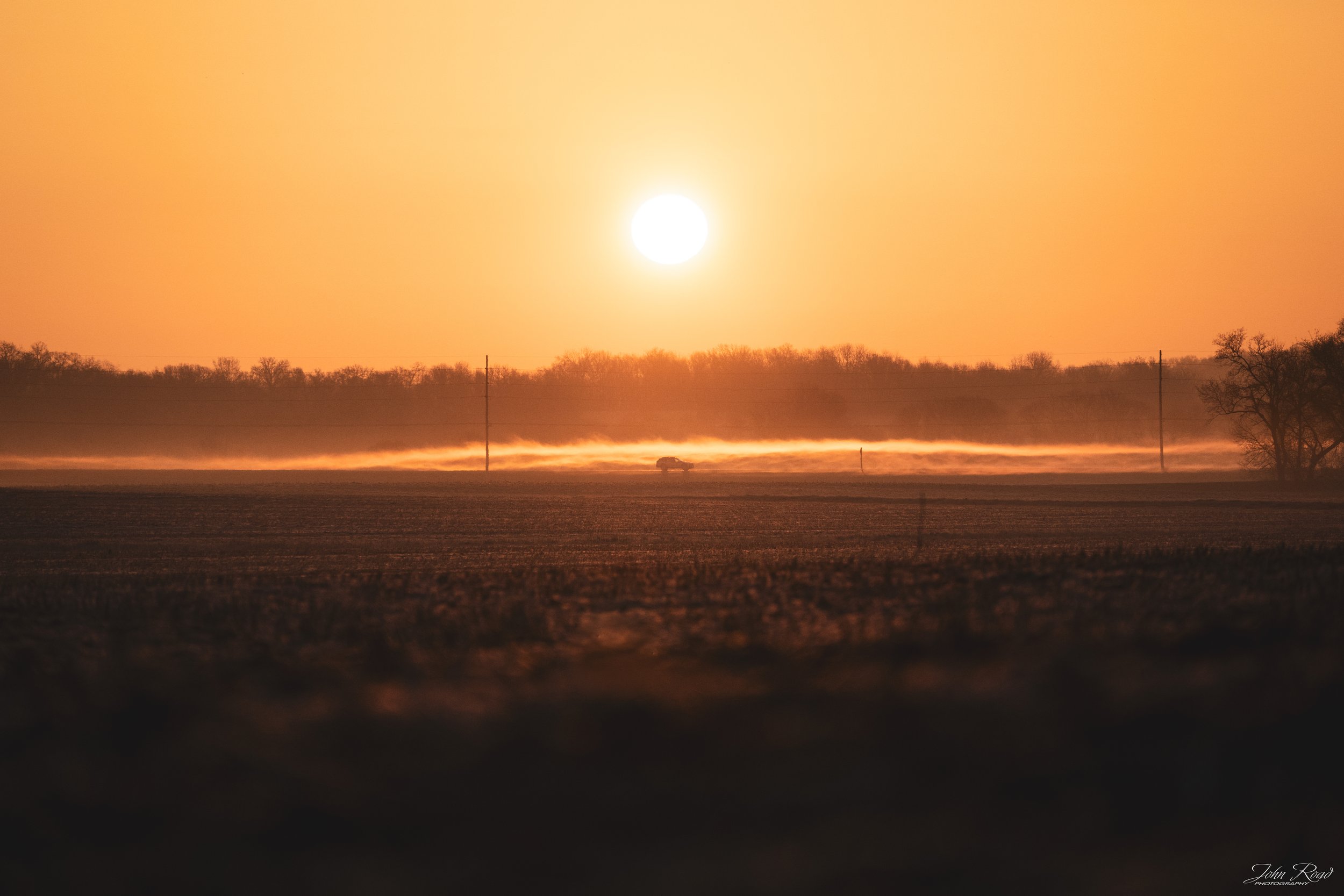 Warm sunrise over a rural field with low fog drifting across the horizon and a distant tree line
