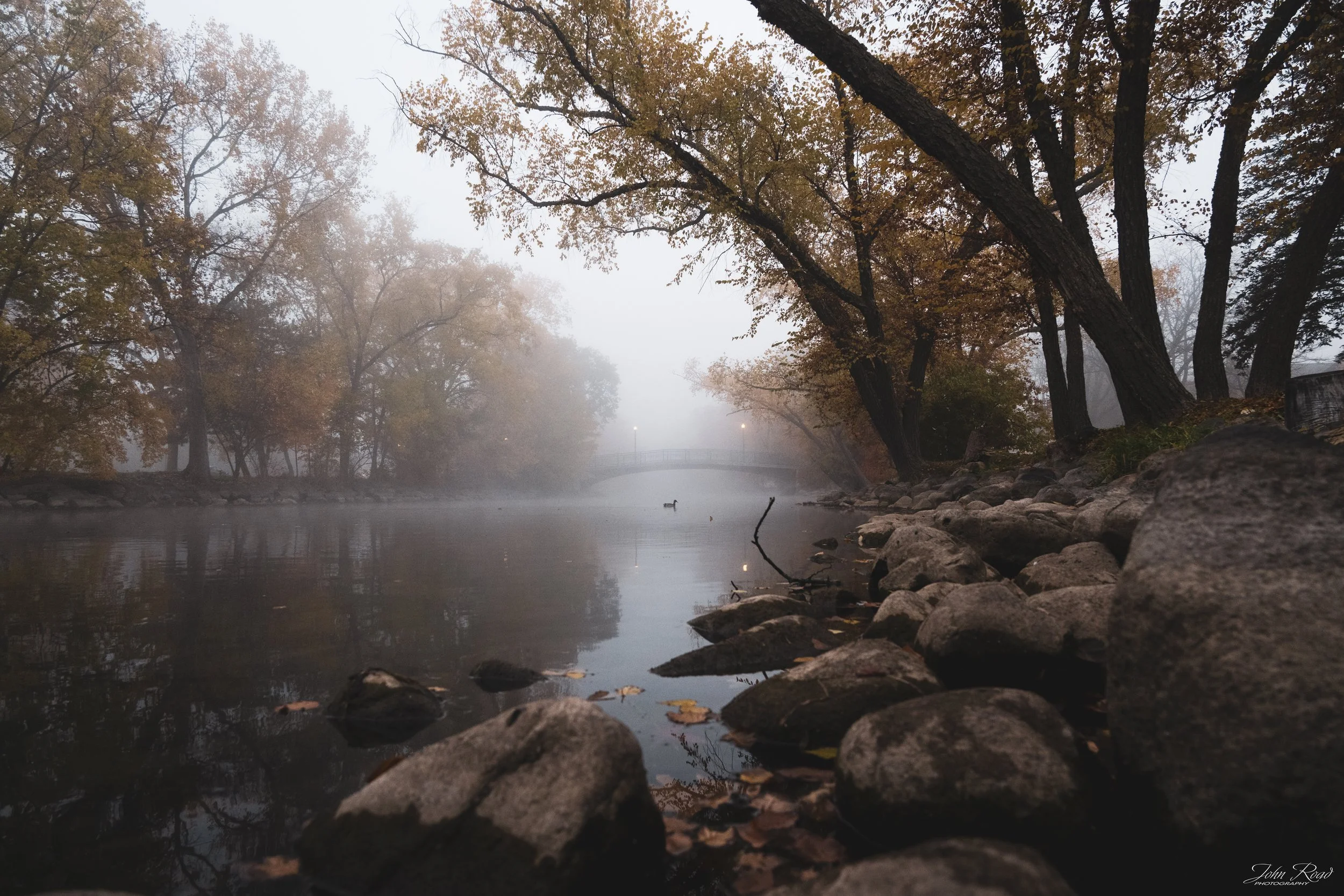 Fine art photograph of stones by a foggy riverbank, soft mist and calm reflections captured by John Road.