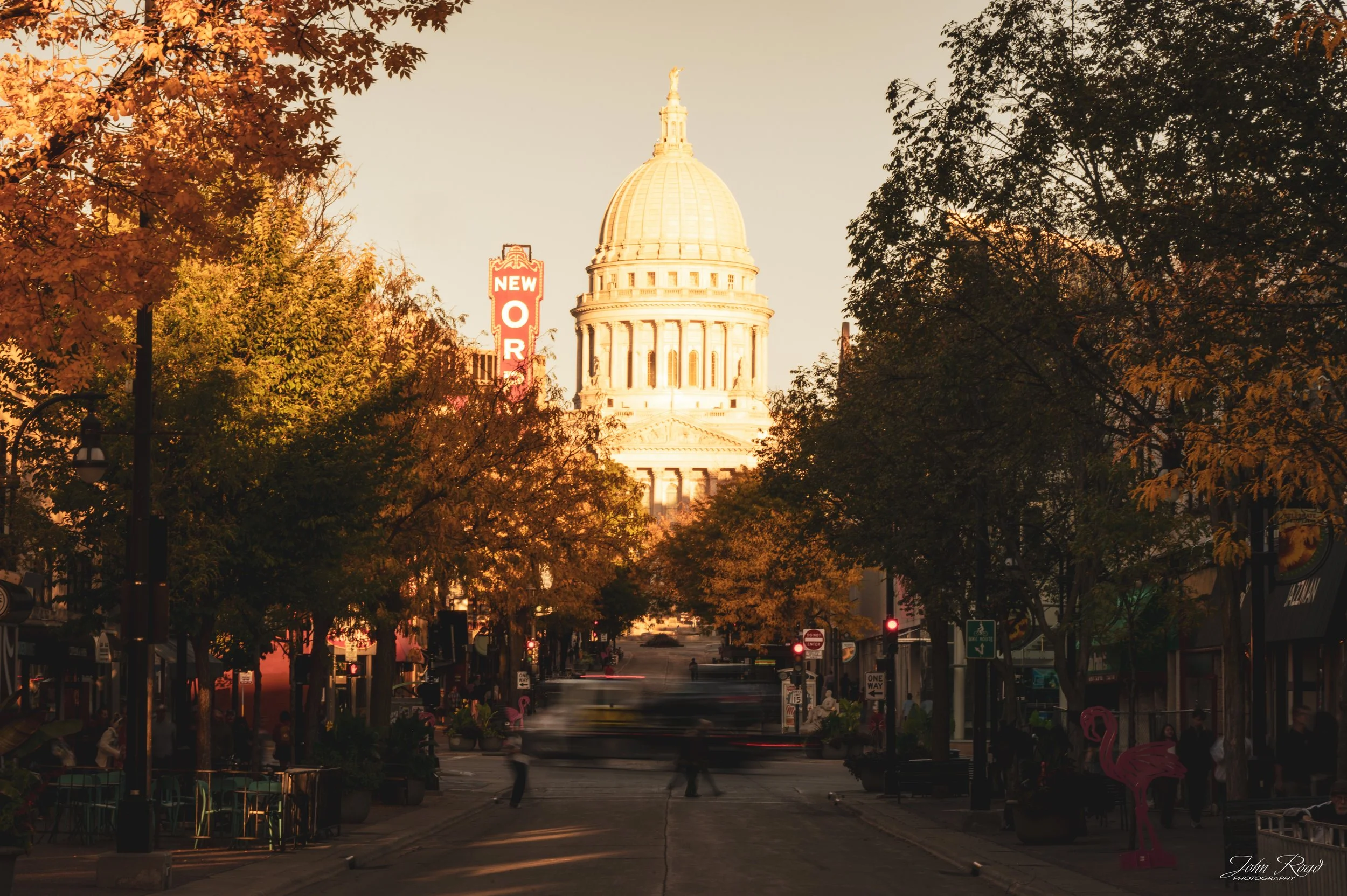 The Wisconsin State Capitol illuminated by warm evening light, framed by autumn trees and historic buildings along a downtown Madison street.