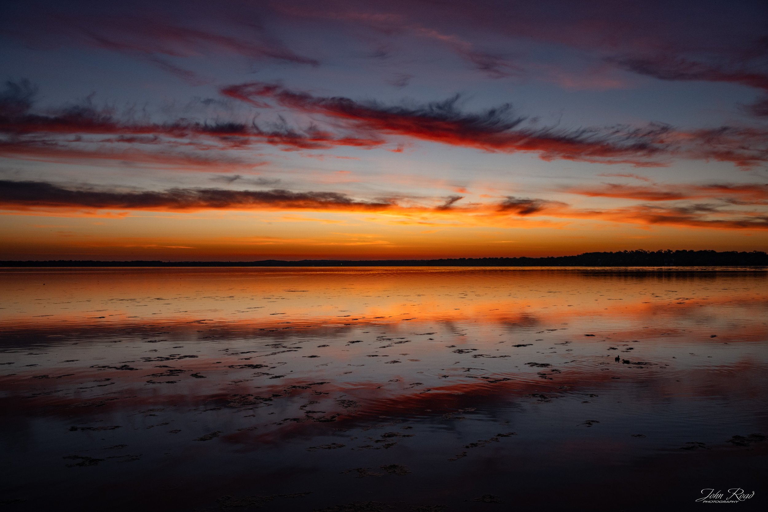 Fire on the Water captures a fleeting moment when the morning sky ignites with color and reflects across the still surface of the lake. Rich reds, oranges, and deep blues create a dramatic contrast, transforming a quiet sunrise into a scene of light,