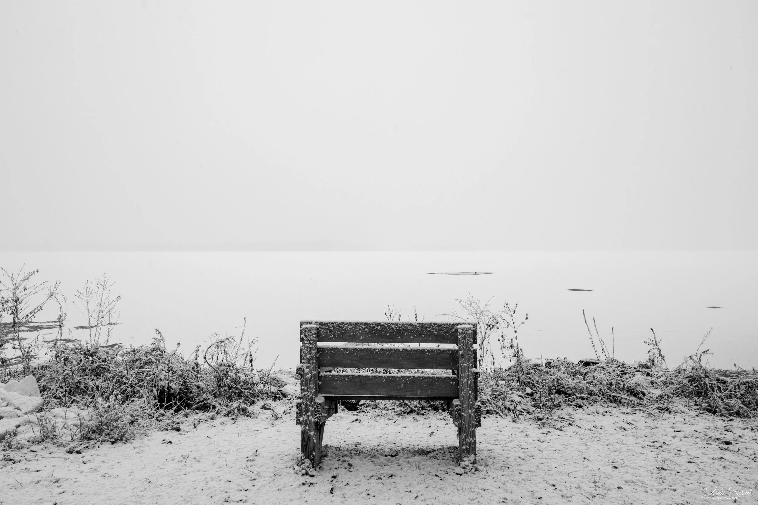 Black and white photo of a lone bench facing a frozen lake in winter