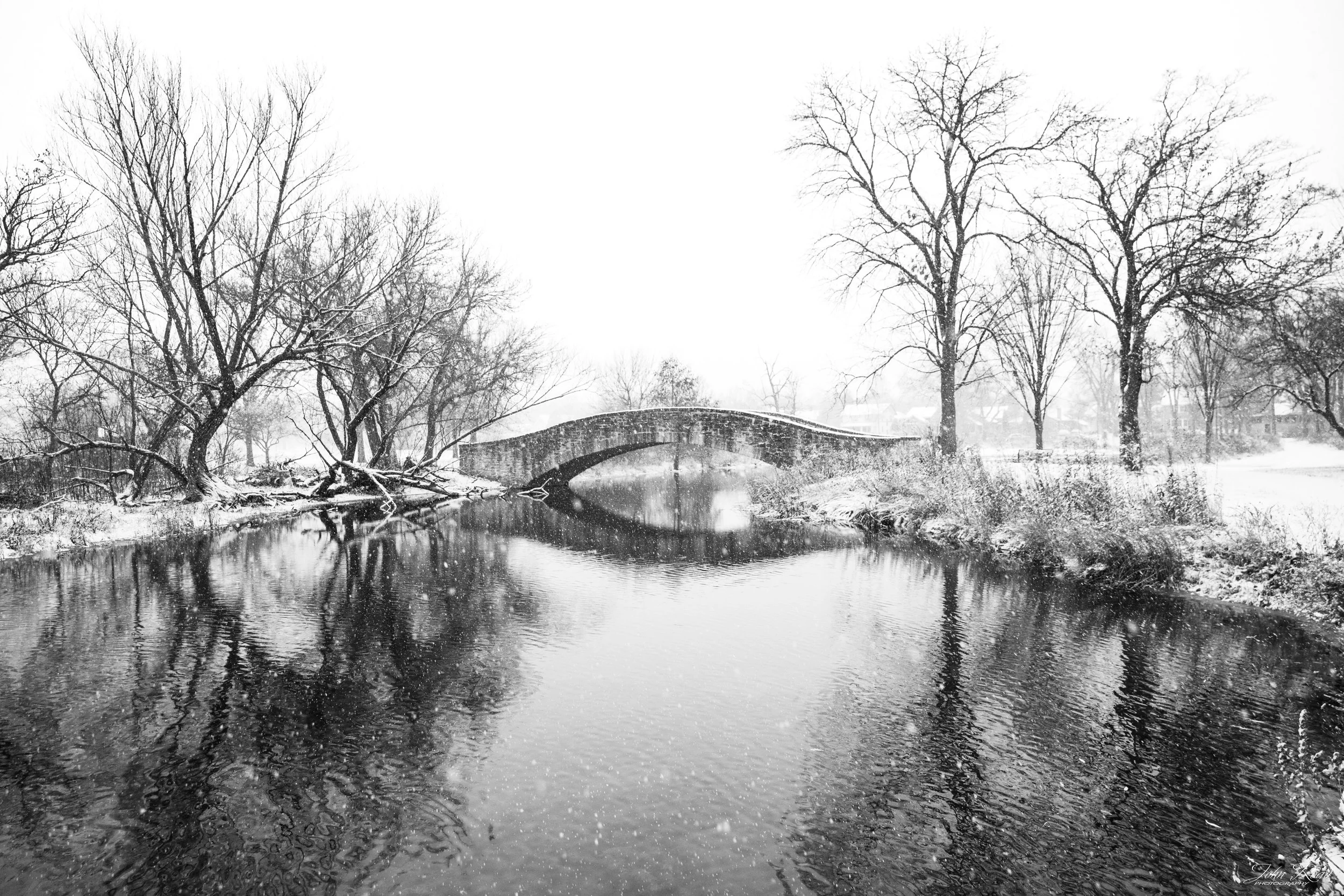 Curved bridge reflected in a winter river with falling snow and bare trees.