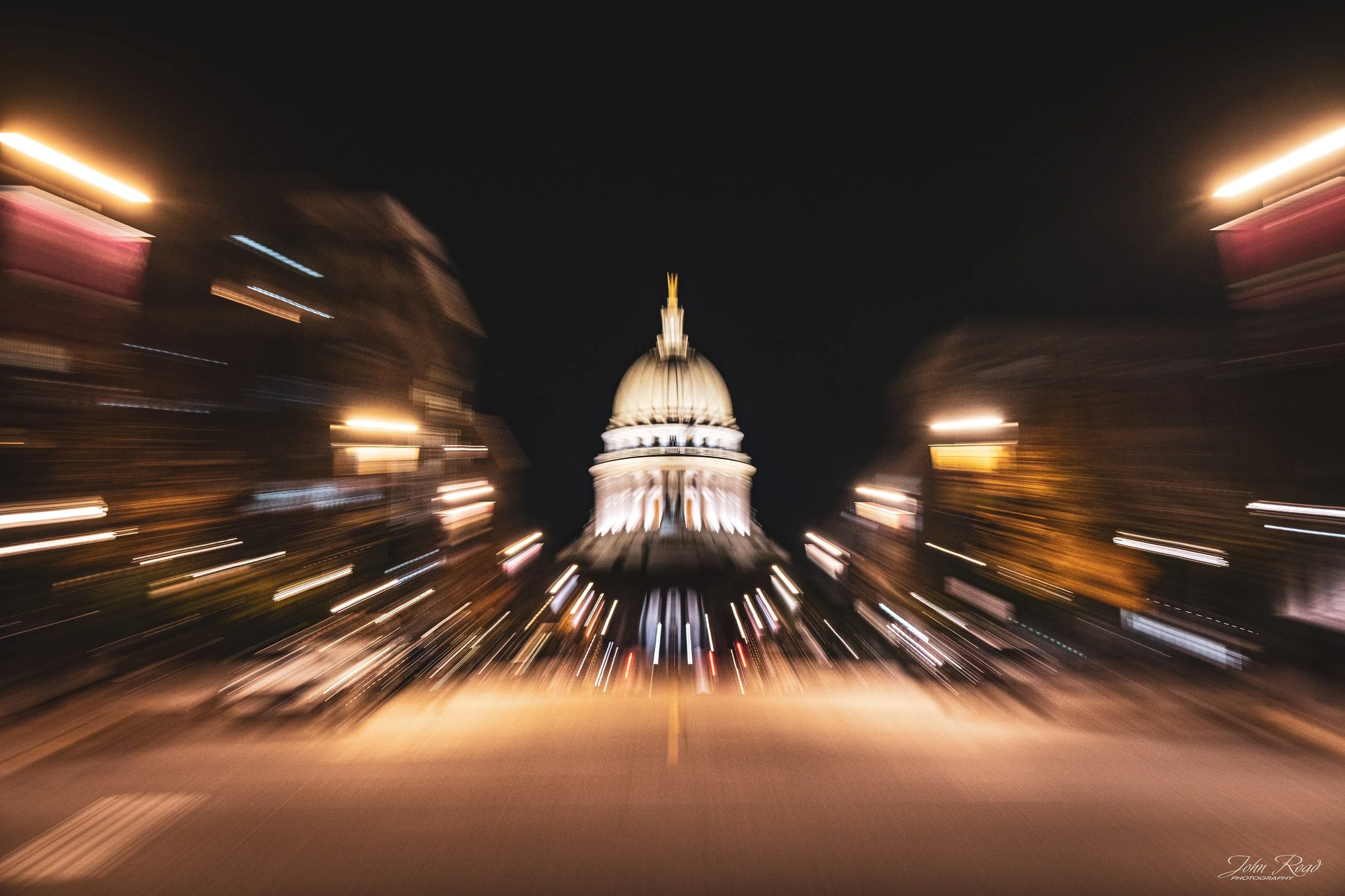 Warm golden light trails and radial motion blur surround the Wisconsin State Capitol and downtown street at night in Madison, Wisconsin.