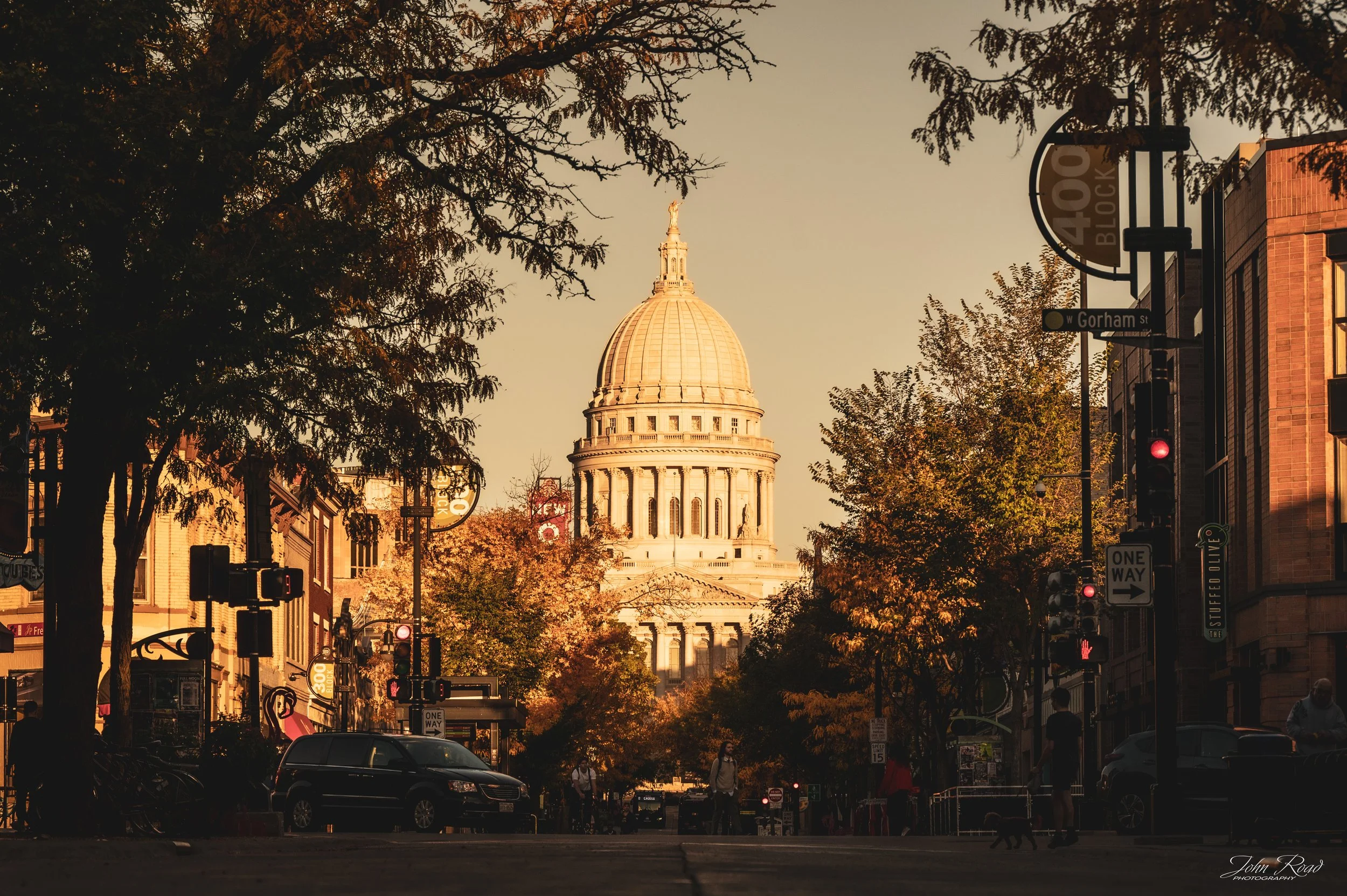 The golden dome of the Wisconsin State Capitol seen through autumn trees along a downtown Madison street in warm evening light.