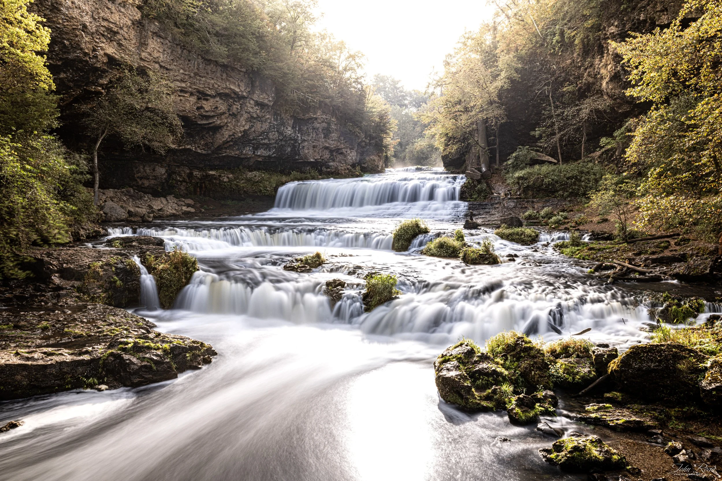 Waterfall at Willow River State Park, Wisconsin, captured by John Road with golden light illuminating flowing cascades and mossy rocks.