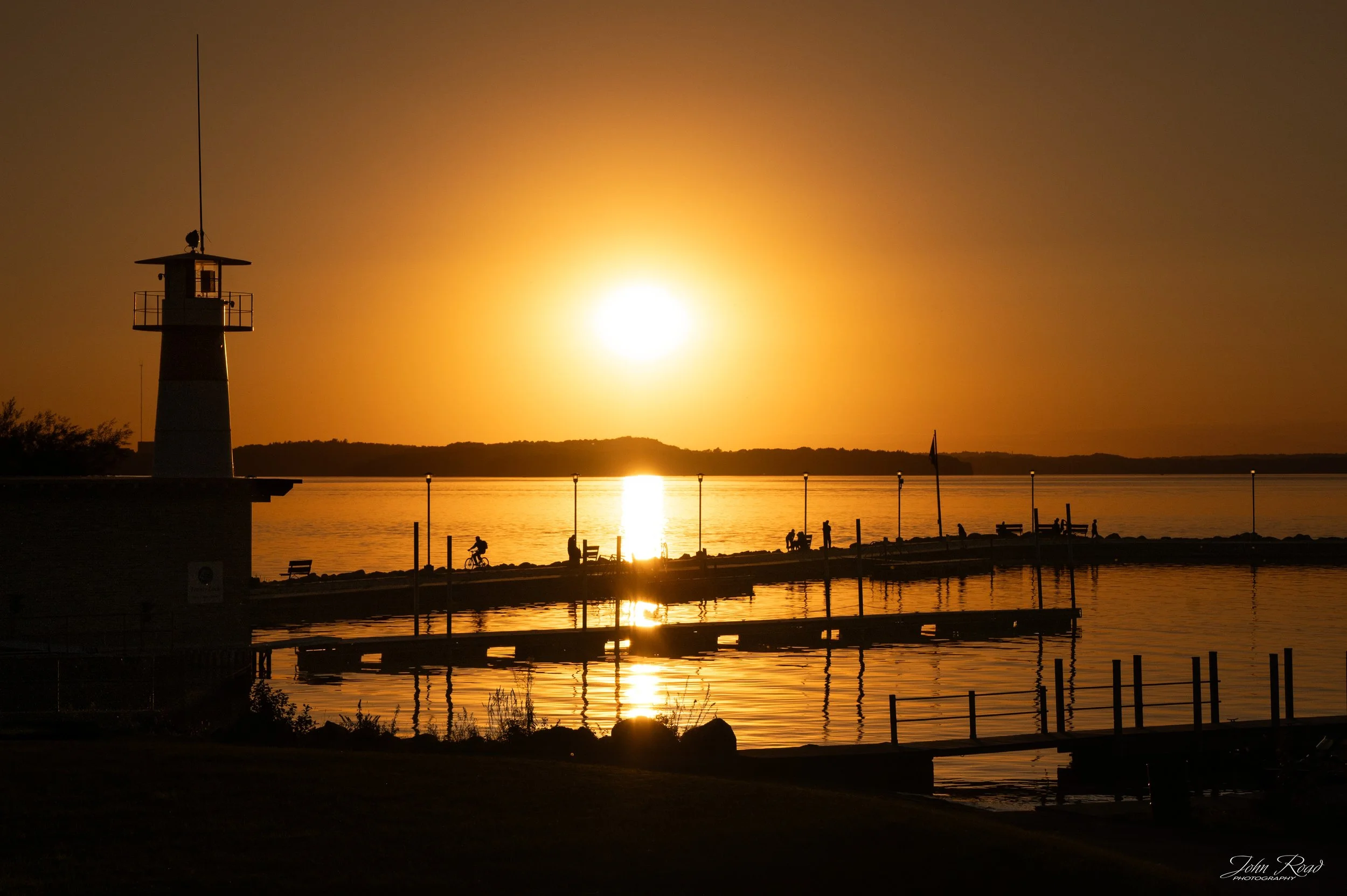 A lighthouse and wooden pier stand in silhouette against the setting sun, with a bright golden reflection stretching across the water.