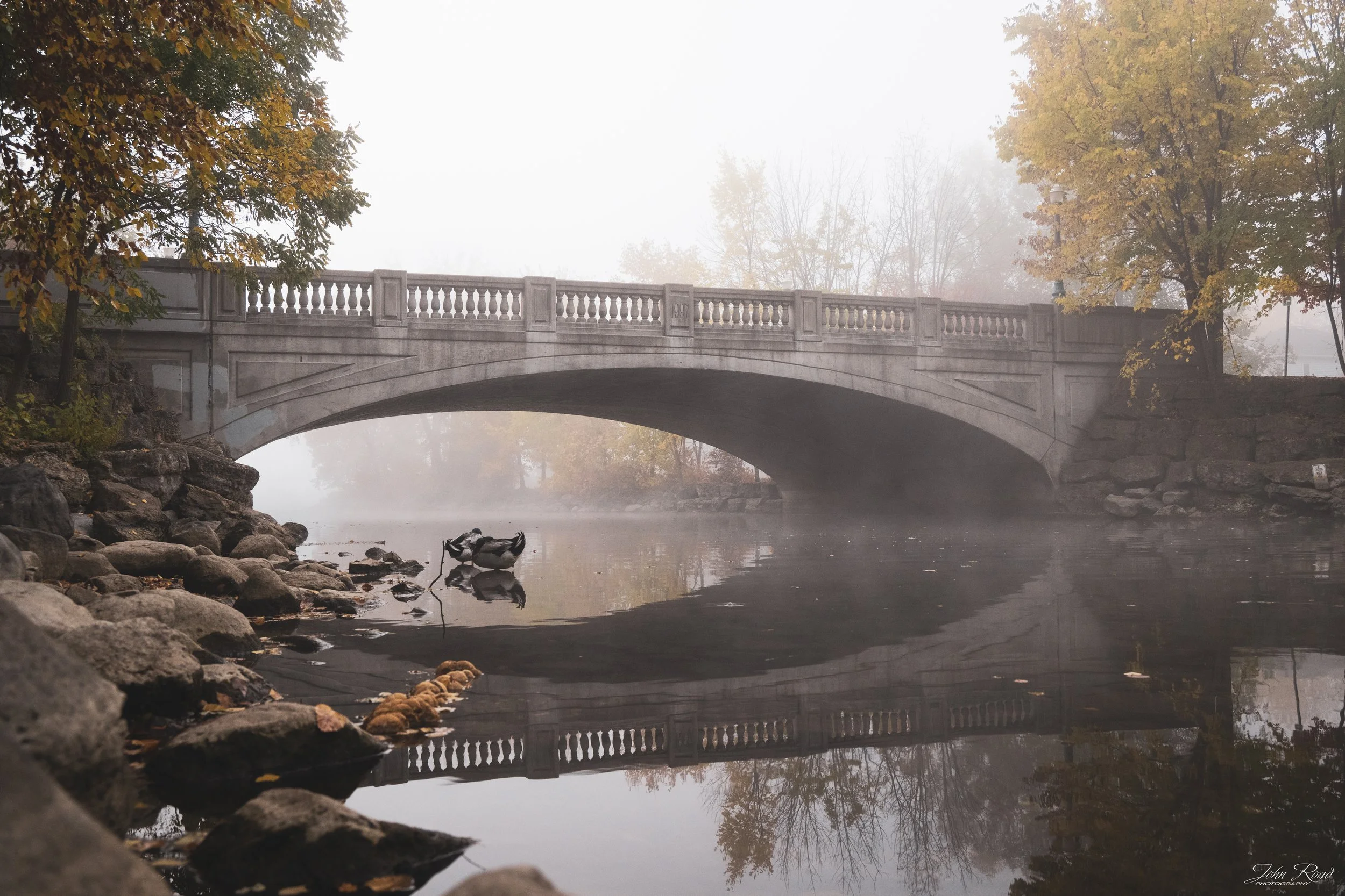Fine art landscape photograph of a bridge surrounded by fog and reflected in still water, captured by John Road.