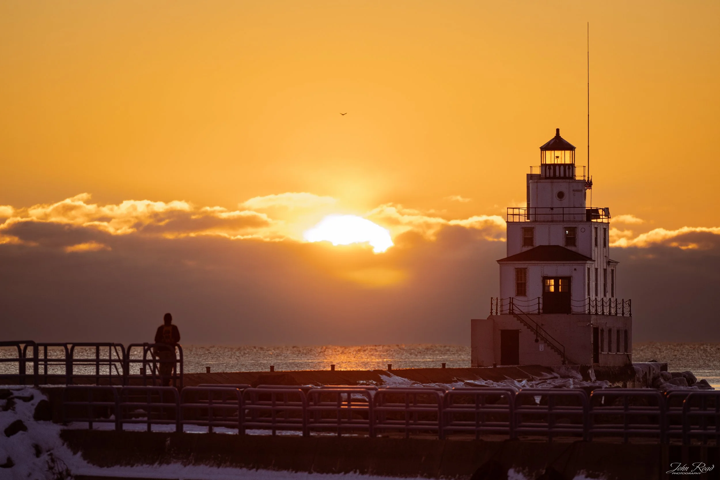 Person standing near Manitowoc lighthouse during winter solstice sunrise on Lake Michigan, warm golden light and snowy pier, photograph by John Road.