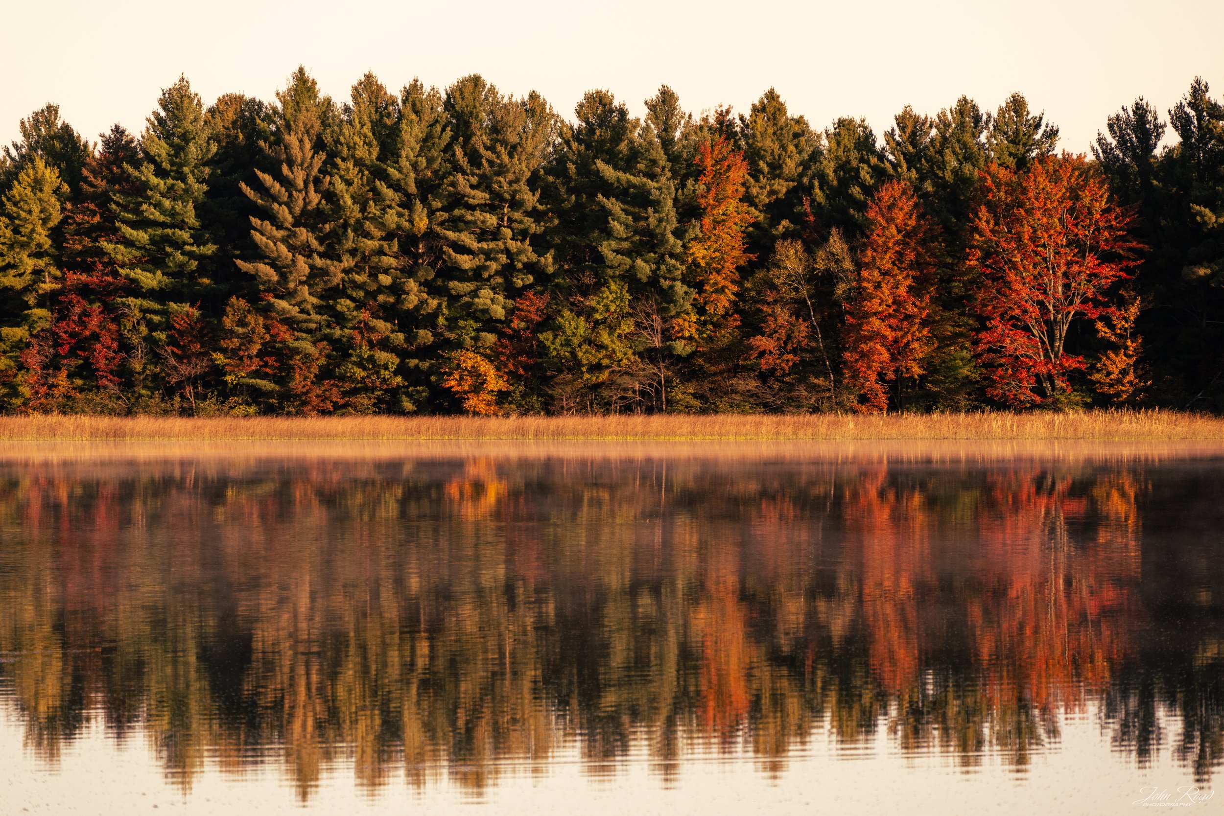 Reflection of autumn forest on still lake water with red and green trees, photographed in Wisconsin by John Road.