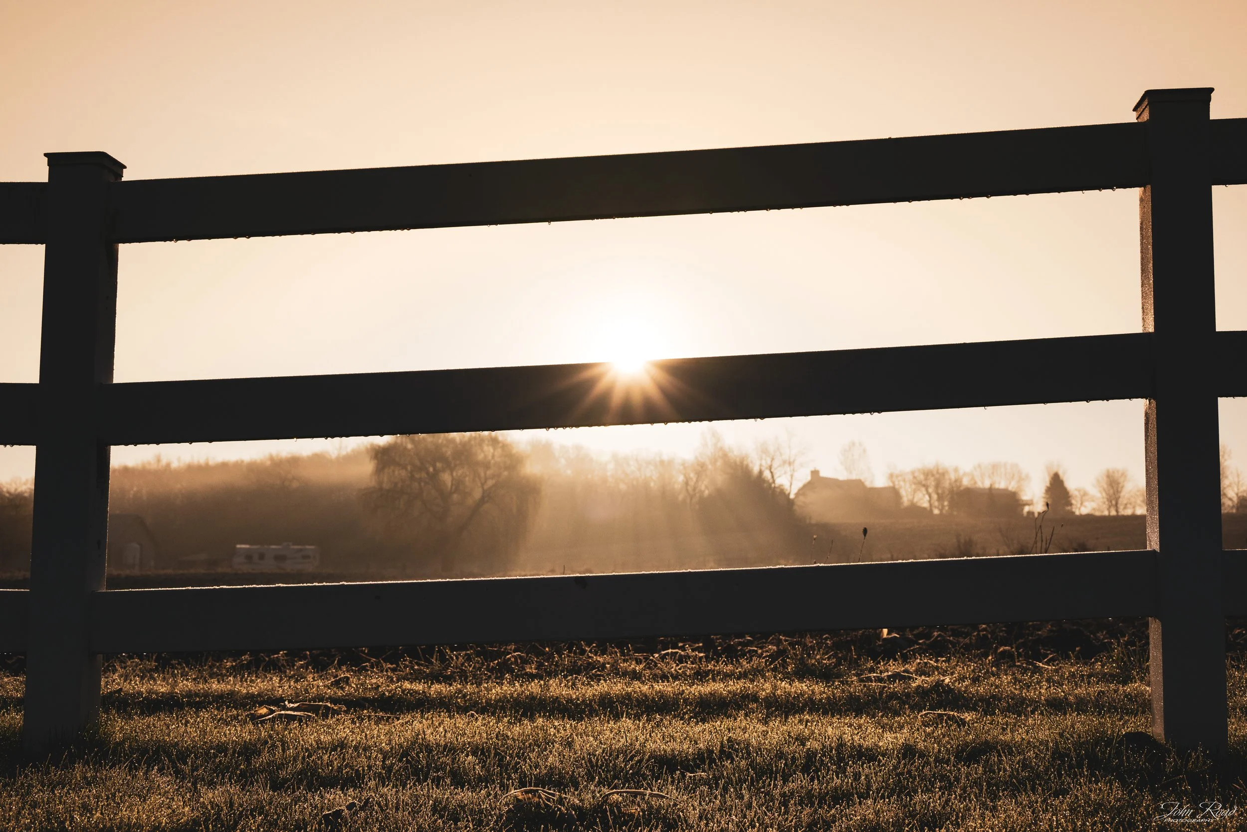 Golden sunrise shining through a wooden fence over a rural field