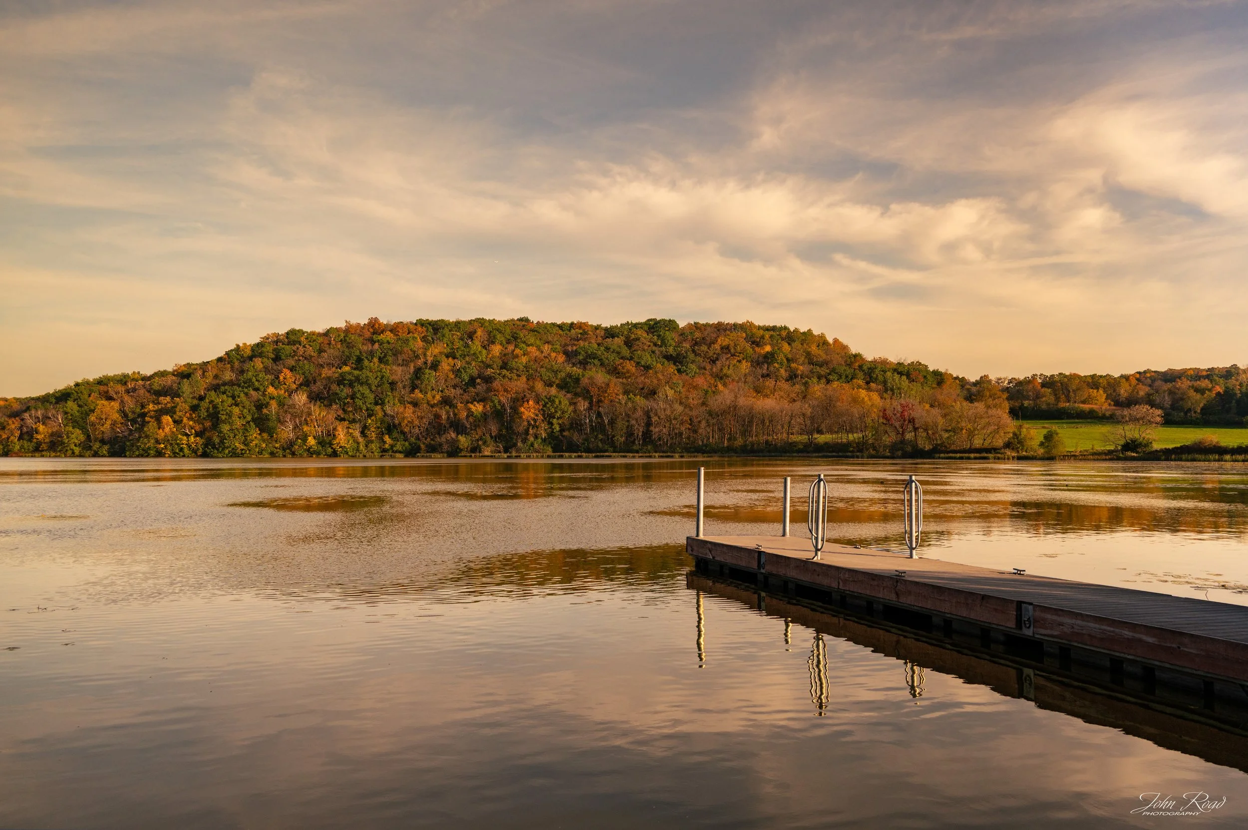 A wooden dock extends into the calm water of Indian Lake, Wisconsin, surrounded by colorful autumn foliage.