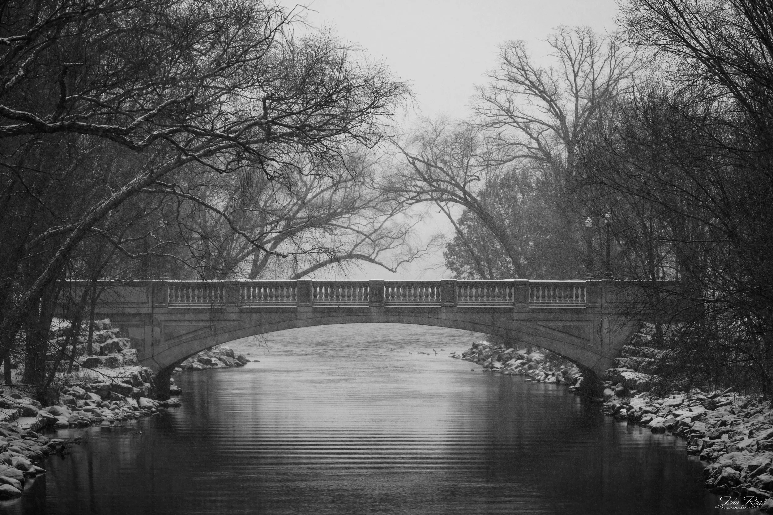 Snowy black-and-white photo of a historic bridge over a quiet river surrounded by winter trees.