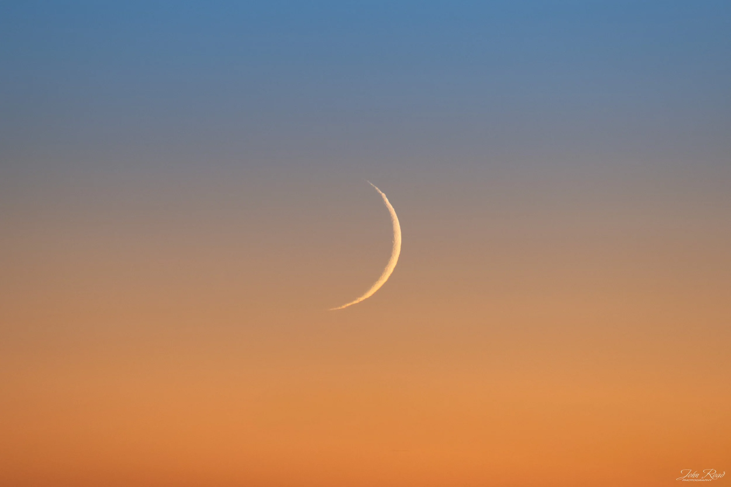 Crescent moon above a colorful sunset sky fading from orange to blue, photographed in Wisconsin by John Road.