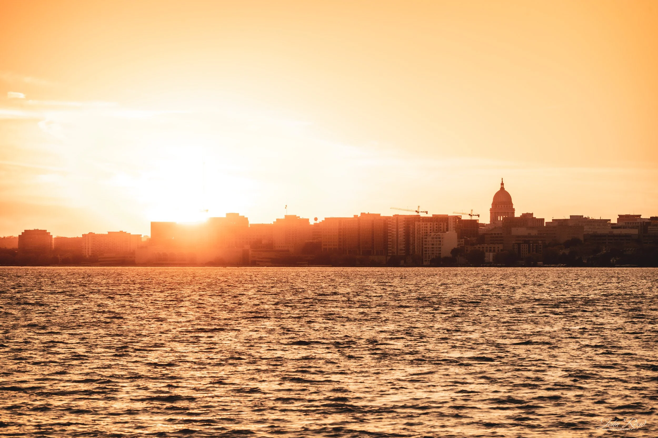 The Madison skyline and Wisconsin State Capitol appear in silhouette across the lake beneath an orange and golden evening sky.