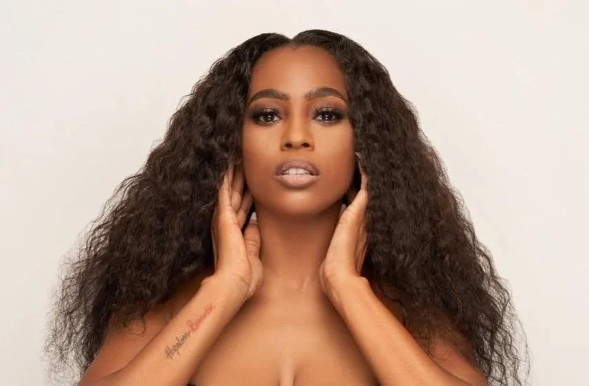 Woman with long curly hair and a tattoo on her forearm, posing with her hands near her face against a neutral background.