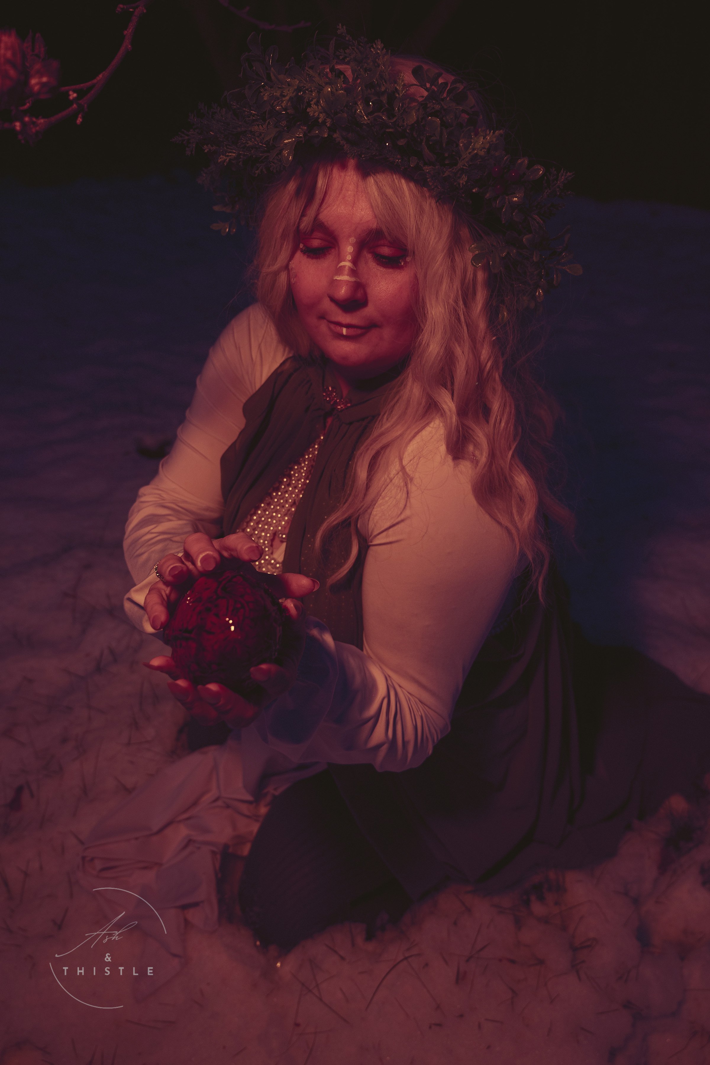 A woman with long wavy blonde hair wearing a floral wreath on her head, holding a red glowing orb while kneeling on a bed of cotton in a dimly lit setting.