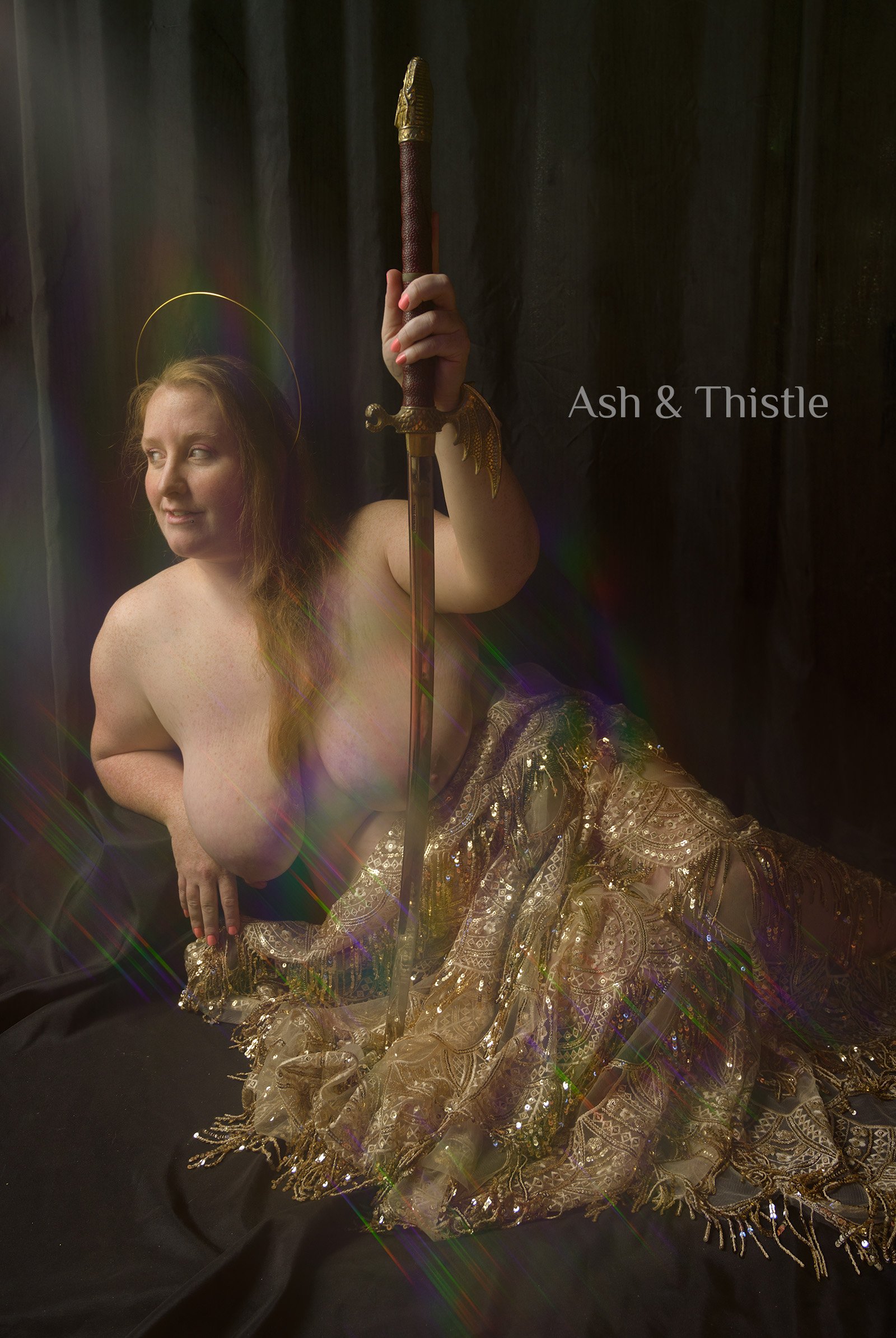 A woman with long red hair and a halo is reclining on the floor. She holds a sword with a decorative hilt. She is topless and wears an ornate, sheer, gold-colored skirt. The background is dark with some rainbow-colored light effects. The text 'Ash & Thistle' is present in the image.